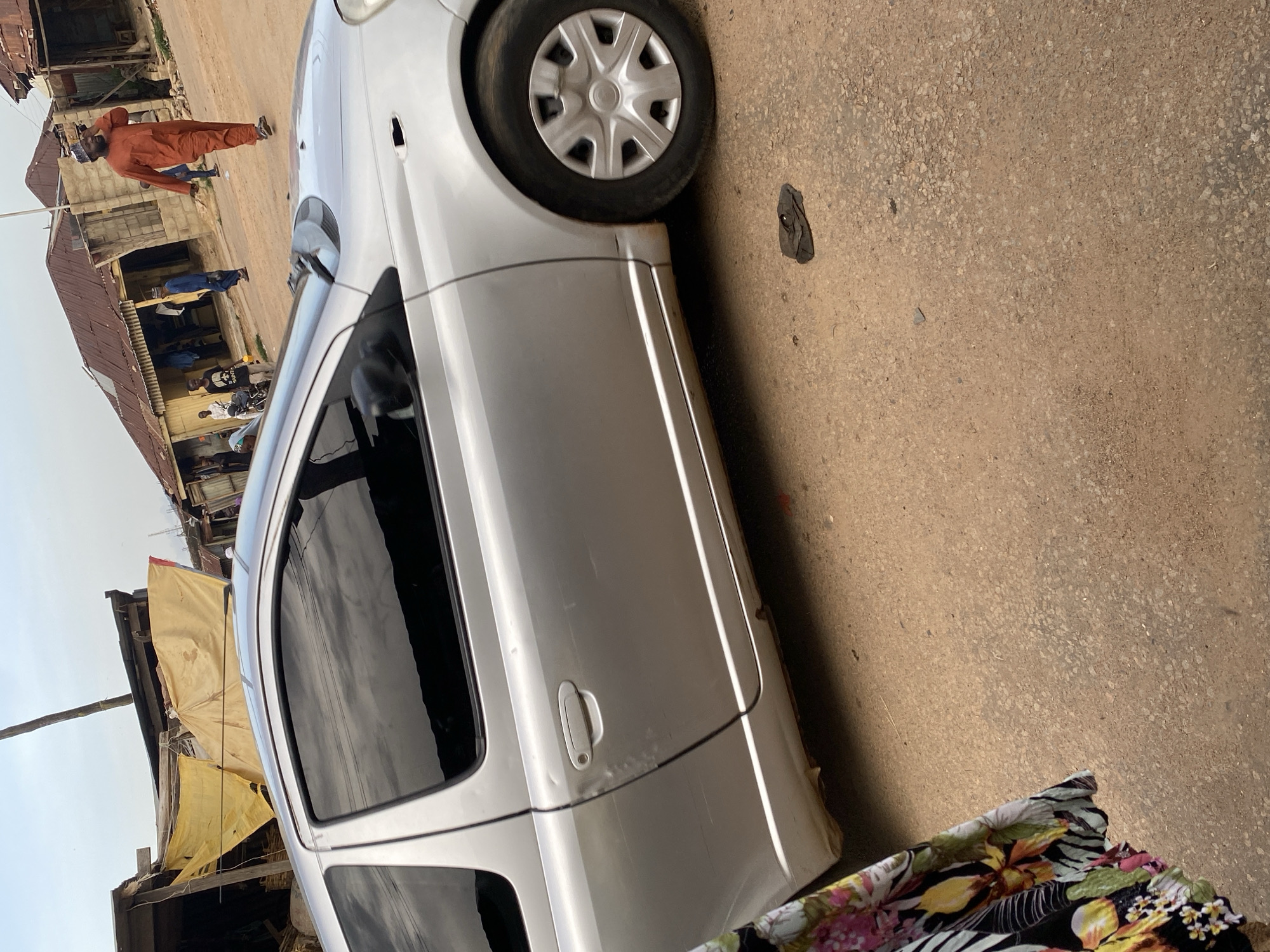 A silver Toyota Yaris is parked on a dirt road in a Nigerian urban environment, likely in Jos, Nigeria. The car is damaged, with visible scratches and dents on its side. A person wearing an orange garment is walking in the background, talking on a mobile phone. Other people and buildings are also visible in the background, suggesting a busy street or marketplace. The ground is covered in dirt and gravel, and there is a piece of dark cloth lying on the ground near the car. The sky is overcast, with signs of an impending rain.