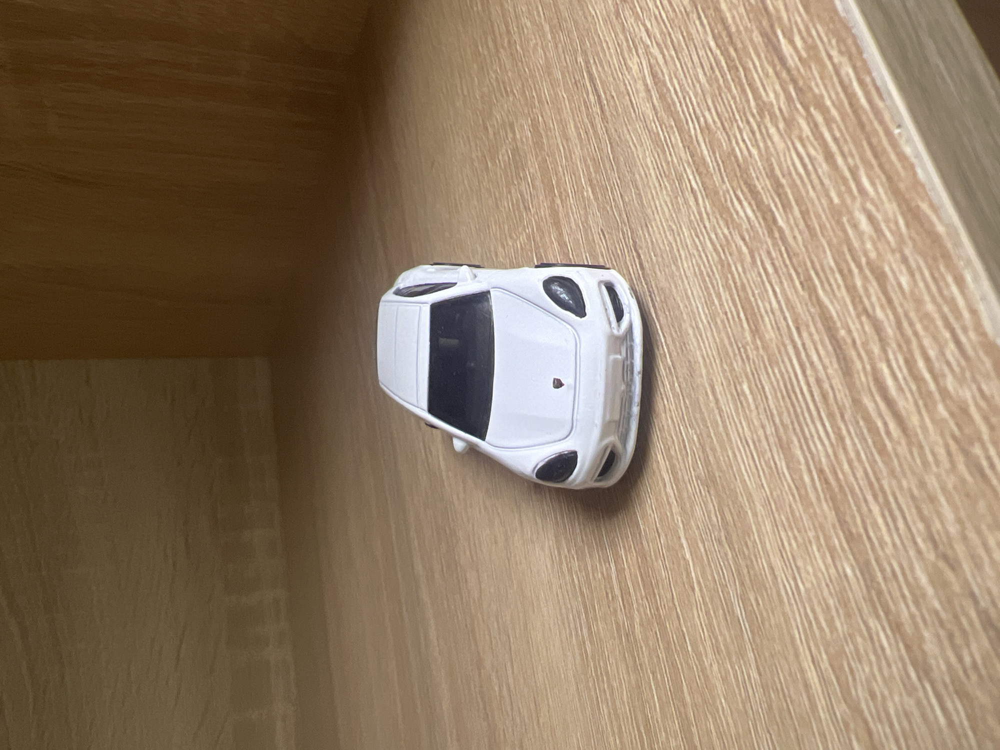 A white toy car, appearing to be a Porsche, is positioned on a light brown wooden shelf. The car is facing the viewer and is centrally located on the shelf. The background consists of the interior of a cabinet or shelf, also made of light brown wood. The lighting suggests an indoor environment, possibly a home or store display. There are no people or discernible actions taking place. The image does not provide any information that would indicate its location in Minsk, Belarus.