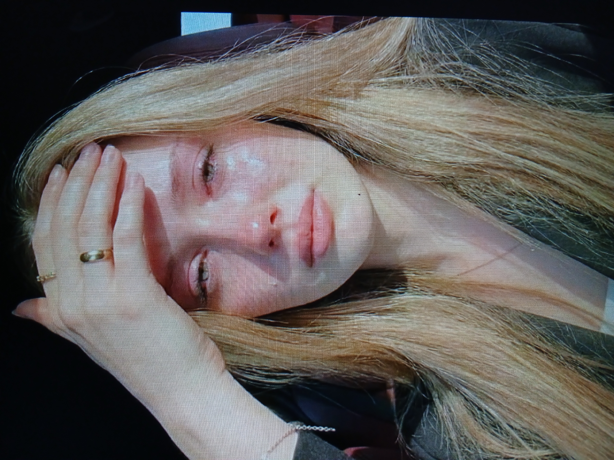 This is a close-up, head and shoulders shot of a young Caucasian female with long, blonde hair. She is outdoors, with her hand resting on her forehead and eyes closed as if in distress or pain. Her eyes are red and watery, and there are visible tear tracks on her cheeks. She is wearing rings on her fingers. The lighting suggests it is daytime. There is no visible indication of the specific location within the city of Dnipro, Ukraine, or any environmental details that would suggest a cityscape.