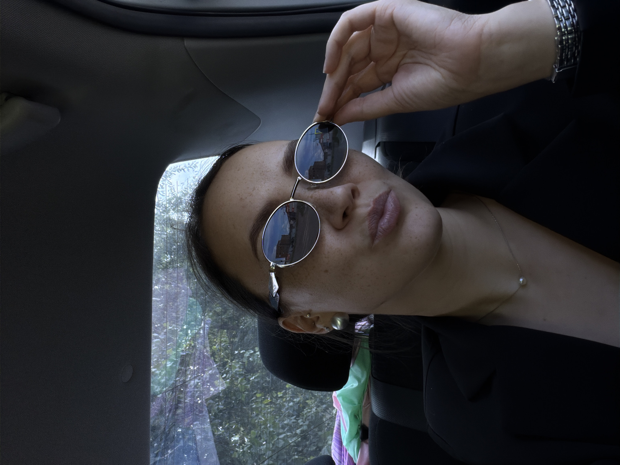A young woman with dark hair pulled back and wearing sunglasses and a black blazer is inside a vehicle, looking at the camera with a playful expression and puckered lips. She is also wearing a pearl necklace and a watch. In the background, through the rear window of the car, trees and buildings are visible, indicating an urban or suburban setting, possibly within the city of Chaikovsky, Russia, given the context provided. The car's interior is dimly lit, with a headrest visible in the foreground.
