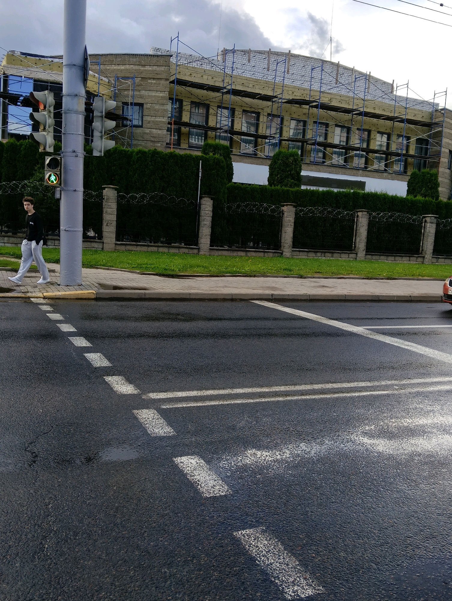 A young man in light gray sweatpants and a dark long-sleeved shirt walks across a pedestrian crossing in Hrodna, Belarus. He is walking from left to right in the image. The street is wet, indicating recent rain. Traffic lights are visible on a pole to the left of the pedestrian, with a green pedestrian signal illuminated. Behind the sidewalk, a fence topped with barbed wire encloses an area with a dense row of green trees. Beyond the trees, a building is under construction, covered in scaffolding and insulation. The sky is overcast.