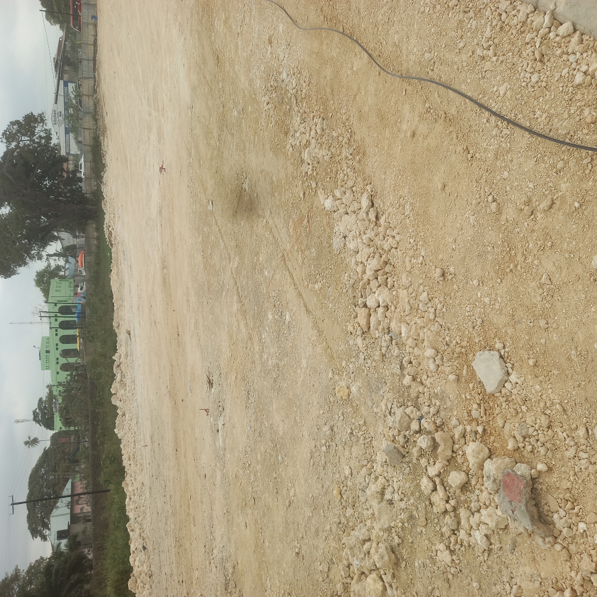 The image depicts a construction site in Bububu, Tanzania, characterized by a vast, uneven expanse of light brown soil and scattered rocks. A dark, coiled cable runs diagonally from the lower right corner across the foreground. In the distance, a row of buildings and trees forms the backdrop. Among these structures, a prominent light green building with arched windows and a white building with signage indicating "Kitchen Bar Solutions" are visible. Construction activity is implied by the presence of orange marker flags and possibly remnants of building materials. The sky is overcast, suggesting a dull or potentially rainy day. There are no people clearly visible in the foreground or mid-ground, but some figures might be present near the distant buildings, though they are too indistinct to be identified. The overall impression is one of an active or recently worked construction area undergoing preparation.
