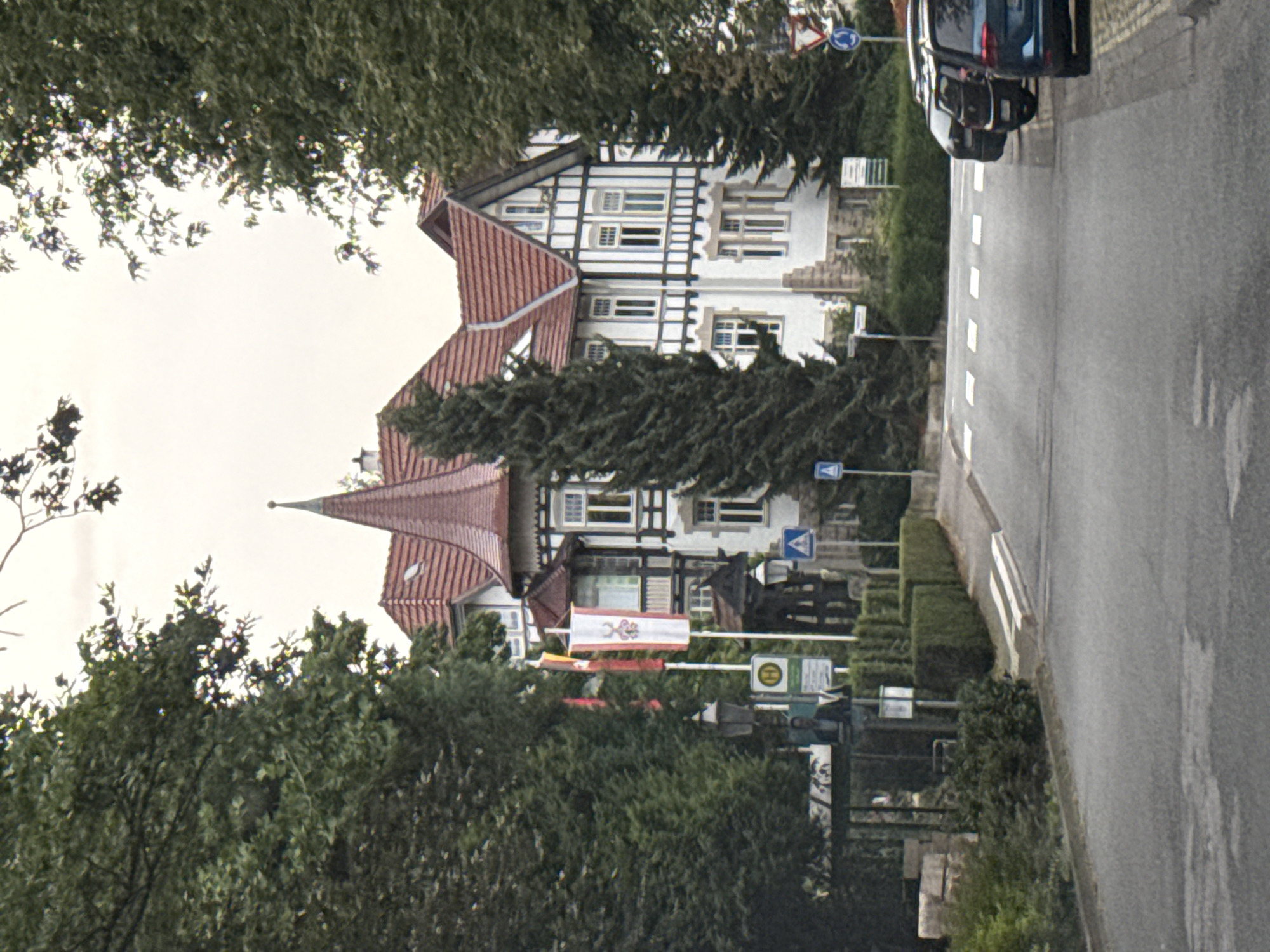 The image captures a street scene in Bad Sachsa, Germany, with a prominent building resembling a villa or historic hotel in the background. The building features a red tiled roof, white walls with dark timber framing, and numerous windows. A tall, conical turret with a red roof is a distinctive architectural element. In front of the building, there are several flags on poles, one of which is a white banner with a red emblem.
To the left of the building, mature trees with lush green foliage overhang the scene, casting shadows. A street runs through the foreground, with a pedestrian crossing marked by white stripes. Several cars are parked on the right side of the street. Traffic signs, including a circular sign with a red border and a blue center, and a triangular sign with a blue border and a white arrow, are visible near the intersection.
On the left side of the street, there is a bus stop shelter with a bench. A person in a suit jacket appears to be standing near the bus stop, partially obscured by the foliage. Other street furniture, such as lampposts and smaller signs, are also present. The overall impression is a well-maintained urban or semi-urban environment, likely a tourist area given the flags and the architectural style of the building. The sky is overcast.