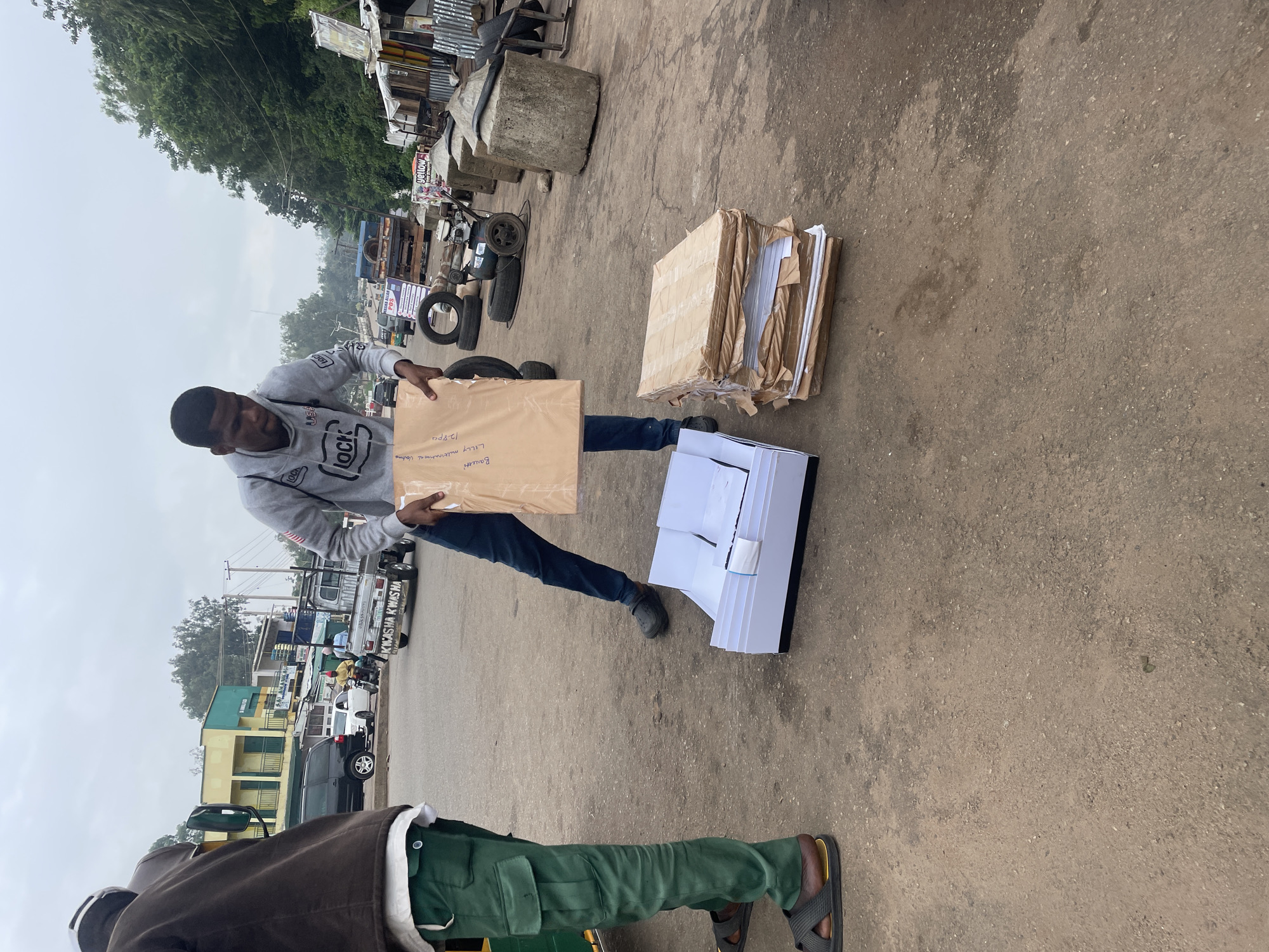 In Jos, Nigeria, two individuals are present in an outdoor setting with a dusty, paved ground. The person on the left, wearing green cargo pants and dark sandals, stands next to a yellow and green tricycle. The person on the right, dressed in a grey hoodie and dark jeans with black clogs, is holding a brown-wrapped package. In front of them, to the right, are stacked brown-wrapped packages and an open white box. The background shows a street with vehicles, buildings, trees, and overhead wires, suggesting an urban environment.