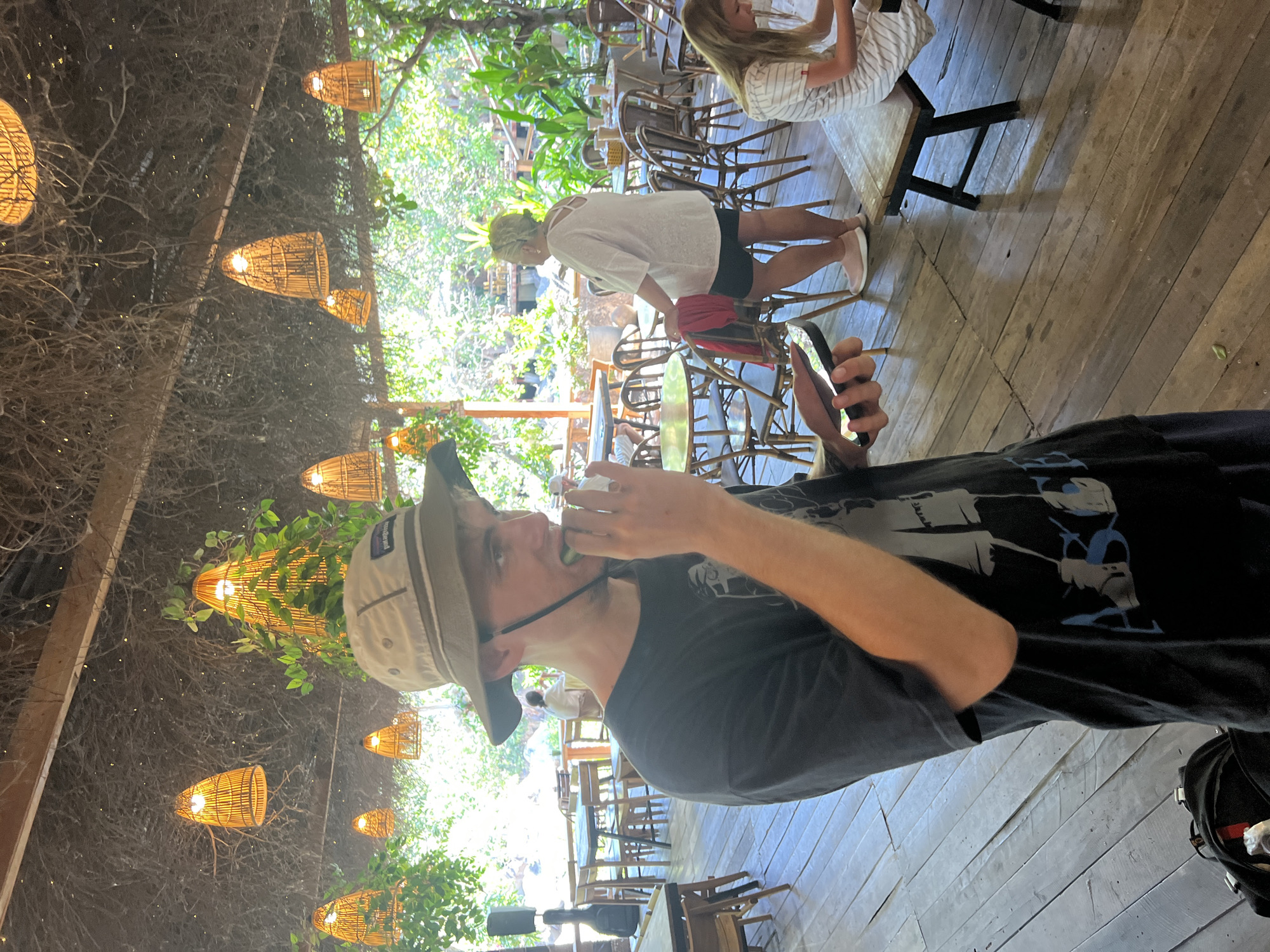 The image depicts an outdoor restaurant or cafe with a thatched roof covered in hanging vines and decorated with numerous woven bamboo pendant lights. Wooden floorboards run throughout the space, with tables and chairs scattered around.
In the foreground, a young person wearing a dark t-shirt and a light-colored bucket hat is captured in profile, holding a smartphone in their left hand and bringing a piece of food, possibly a slice of cucumber, to their mouth with their right hand. They are looking to their right.
In the midground, a woman with blonde hair, wearing a loose white top and dark shorts, stands facing away from the camera, resting her left hand on the back of a chair. Behind her, other diners are visible at tables. To the right, a young girl in a striped dress is seated at a wooden bench, looking down at something on the table.
The background reveals lush green foliage and trees, suggesting a tropical or semi-tropical setting. The overall atmosphere appears relaxed and casual, typical of a cafe or restaurant in a warm climate. While the exact location is not specified in the image, the presence of tropical vegetation and the architectural style of the lighting and roofing could be consistent with a location in Vĩnh Hải, Vietnam, known for its coastal and natural attractions.