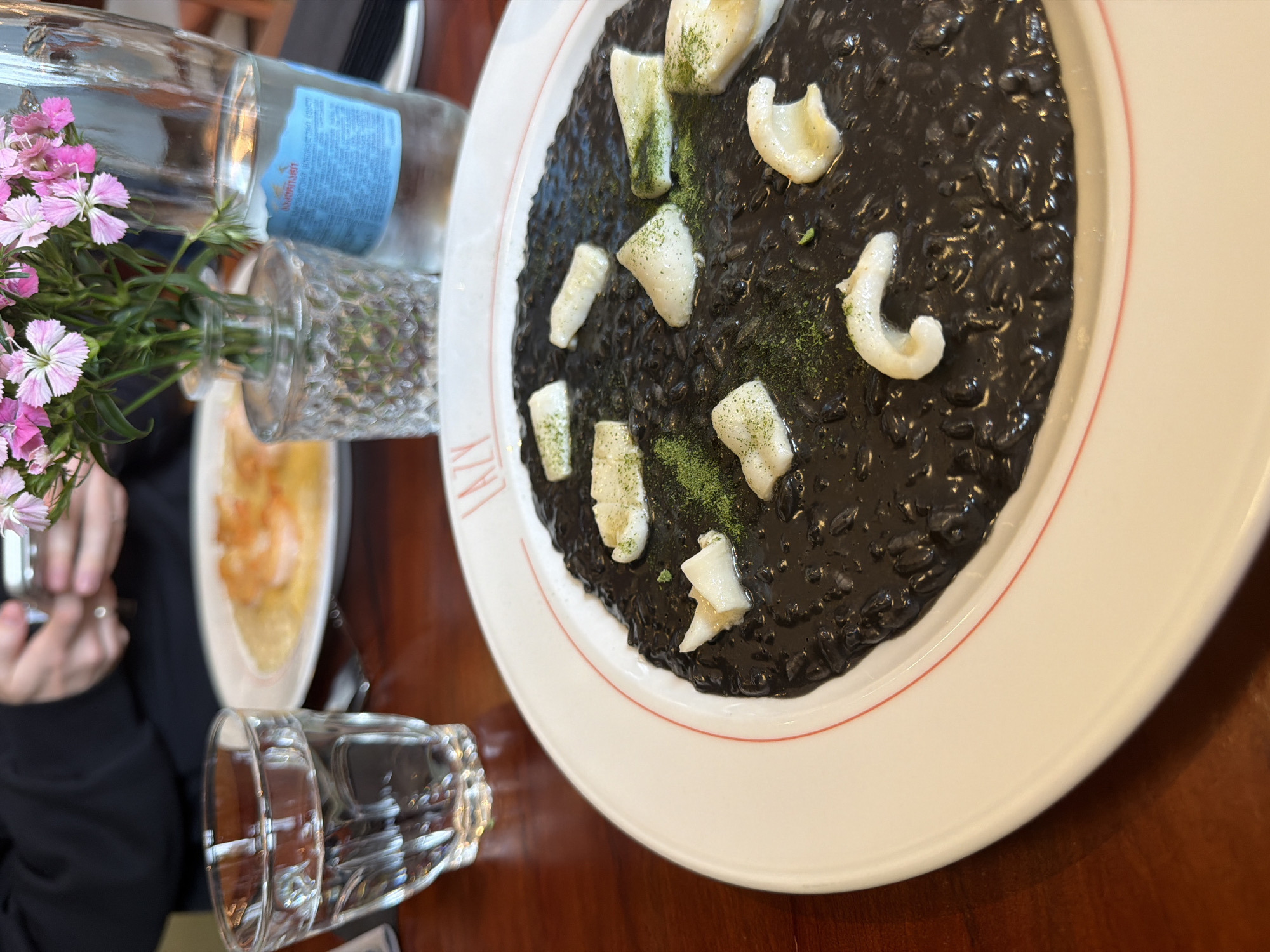 The image depicts a dining table scene, likely in a restaurant. The primary focus is a white plate with a portion of black risotto, garnished with white, possibly seafood, pieces and green specks. The plate has a thin red line around its perimeter.
Next to the plate, there is a glass vase containing pink flowers, along with a bottle of water. An upturned empty glass is near the vase.
In the background, part of another plate with food, seemingly yellow in color, is visible. A person's hand and black clothing can also be seen, slightly blurred, suggesting the diner or photographer. The table has a warm wooden tone.
The letters "LAZY" are printed on the plate with the black risotto. Based on this information and the likelihood of encountering such dishes in that region, the location may be Tbilisi, Georgia.