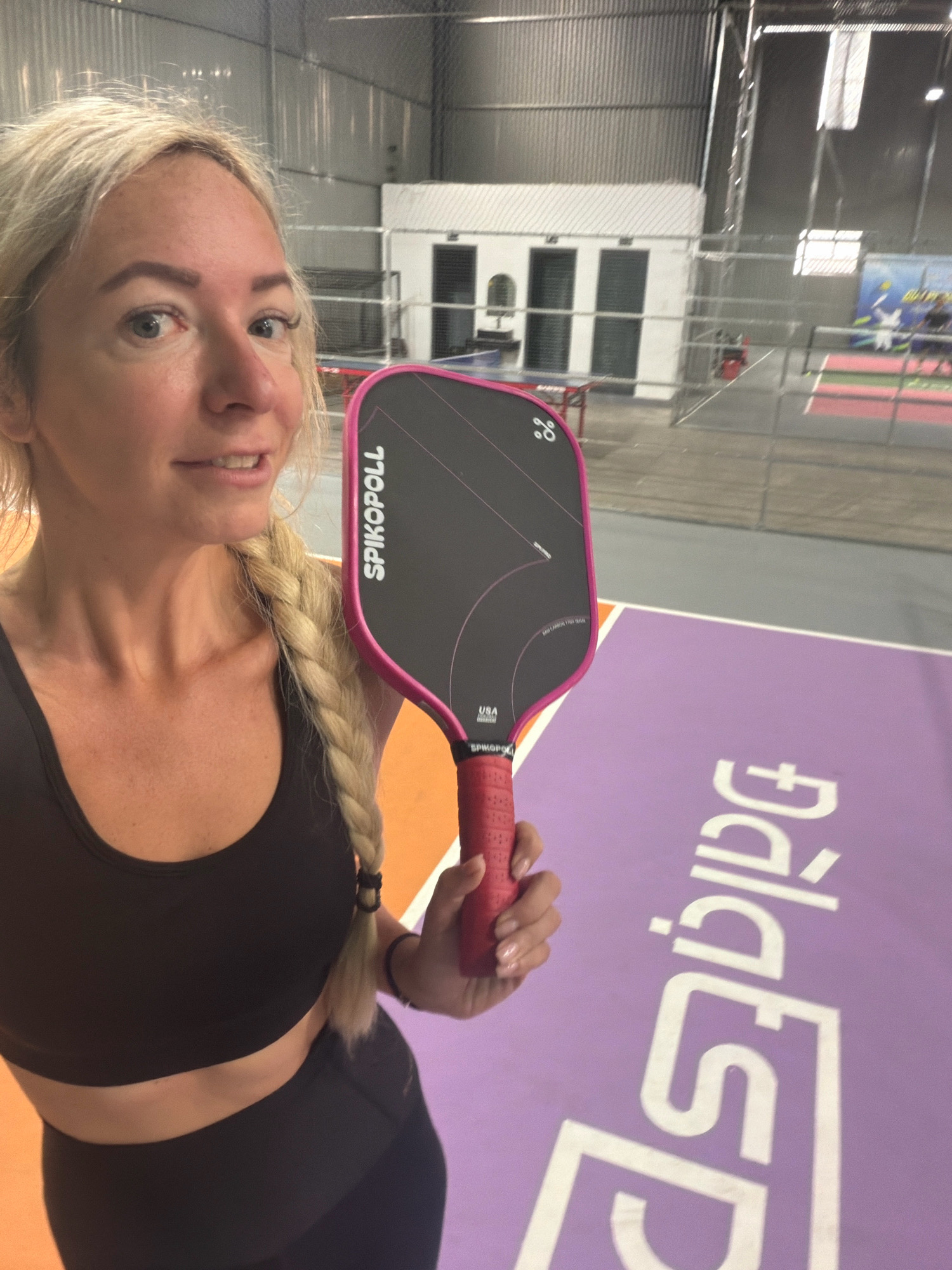 A woman with blonde hair braided over her shoulder holds a pink pickleball paddle. She is wearing a black sports bra and black leggings. The woman is inside what appears to be an indoor sports facility, possibly a pickleball court, as indicated by the court markings on the floor. The court has purple and orange sections with white lettering. In the background, there are netting and other sports equipment. The overall scene suggests an athletic setting.