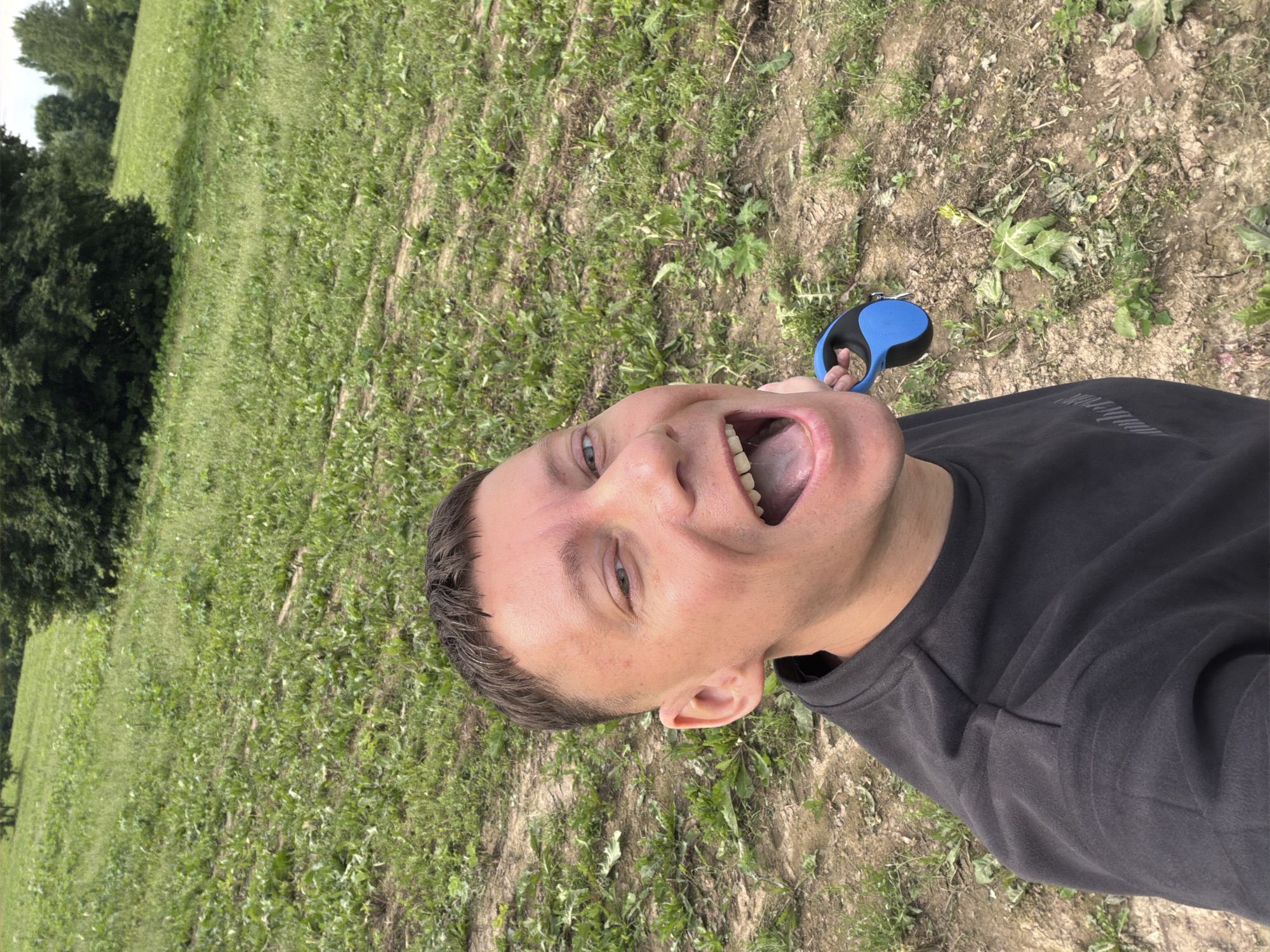 The image shows a close-up, selfie-style shot of a person holding a blue and black retractable dog leash with their right hand, their mouth open in a wide smile, and their eyes looking towards the camera. The person is wearing a dark grey long-sleeved shirt. The background consists of a grassy, uneven terrain with some bare patches of dirt and scattered vegetation. In the distance, there are trees. The scene appears to be outdoors, possibly in a park or a natural area. The lighting suggests it is daytime. There is no visible indication of the specific location within Moscow, Russia.