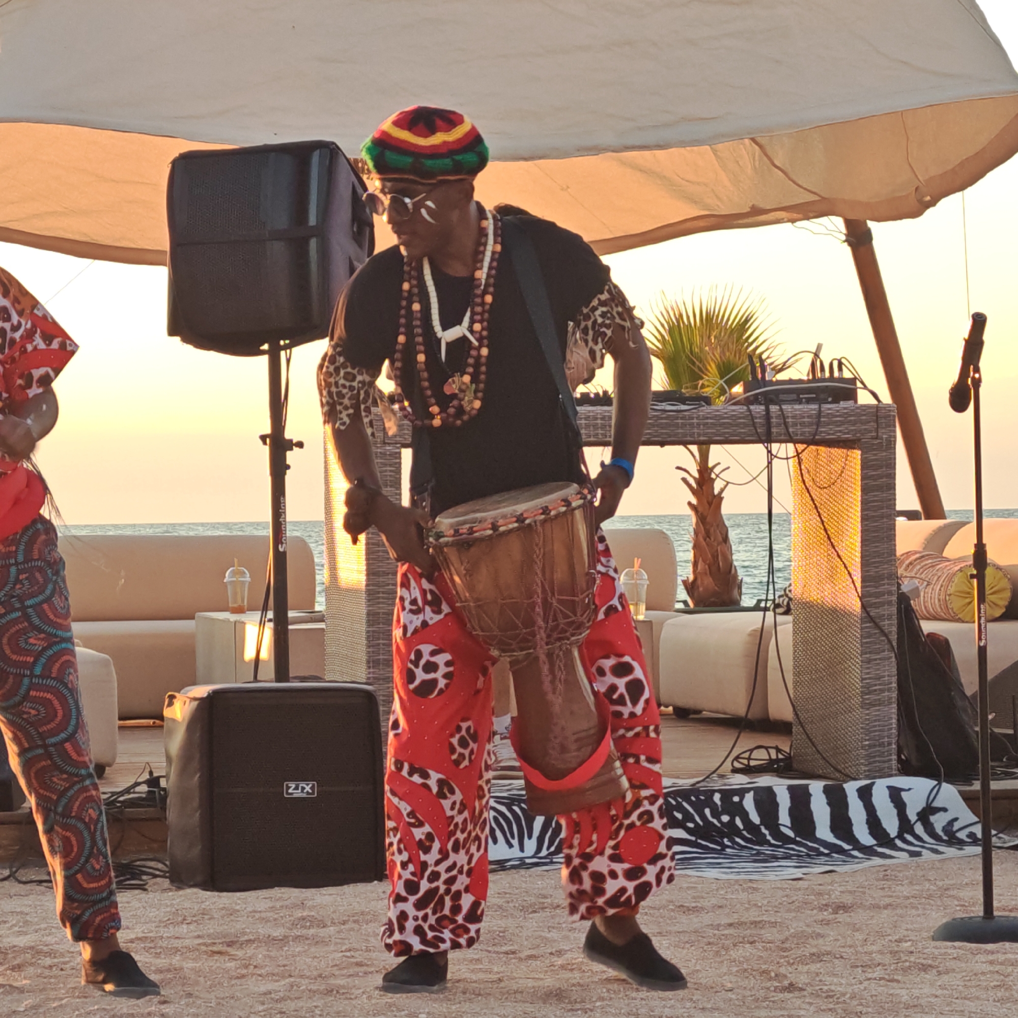 A man, appearing to be of African descent, is playing a djembe drum on a sandy surface. He is wearing a colorful, knitted hat with red, yellow, and green stripes, sunglasses, and a black t-shirt. Around his neck, he wears several layered necklaces made of beads and a white pendant. His arms are adorned with leopard-print armbands, and he is wearing brightly patterned red pants with white and black animalistic prints. He is standing on a beach, with the sea visible in the background. Behind him, there are lounge chairs and a table with DJ equipment. A large speaker is positioned to his left, and a microphone stand is to his right. The overall scene suggests an outdoor performance or event. The sky is illuminated by a sunset, casting a warm glow over the environment. The location is identified as Мысовское сельское поселение, Ukraine.