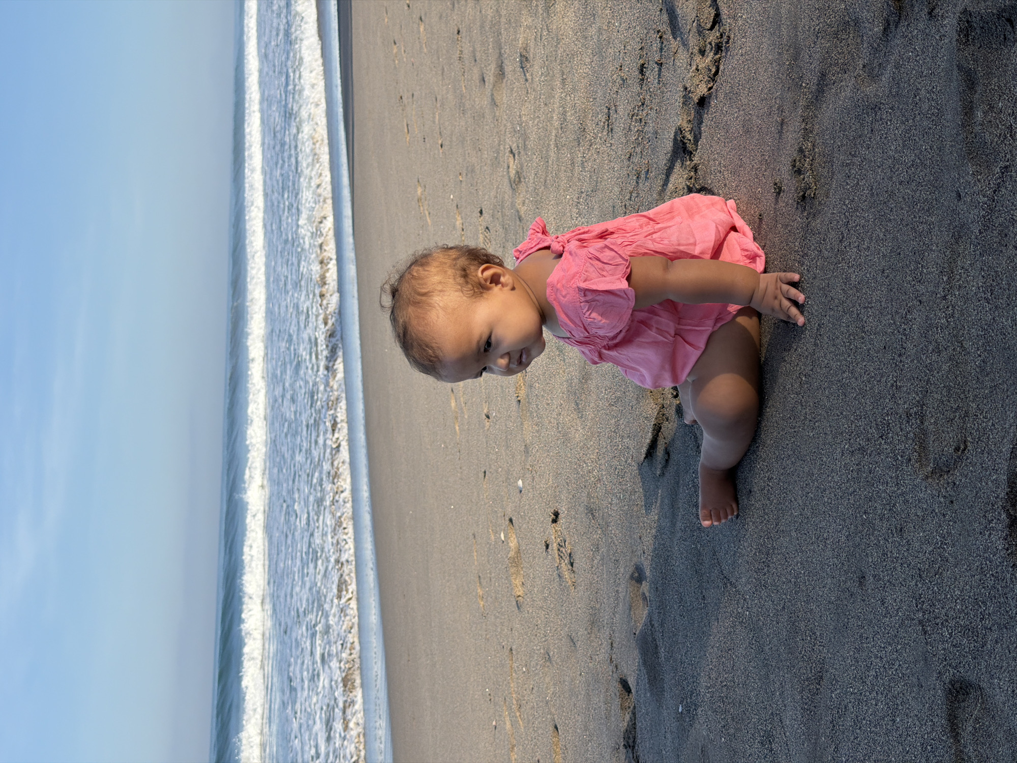 An infant sits on a sandy beach, facing left, with the ocean and its waves in the background. The infant is wearing a pink romper and has dark hair and skin. The sand is dark gray, with some lighter patches and footprints. The water is a vibrant blue-green, with white foam on the waves. The sky is a clear, light blue. The scene is set in North Kuta, Indonesia, likely on a sunny day.