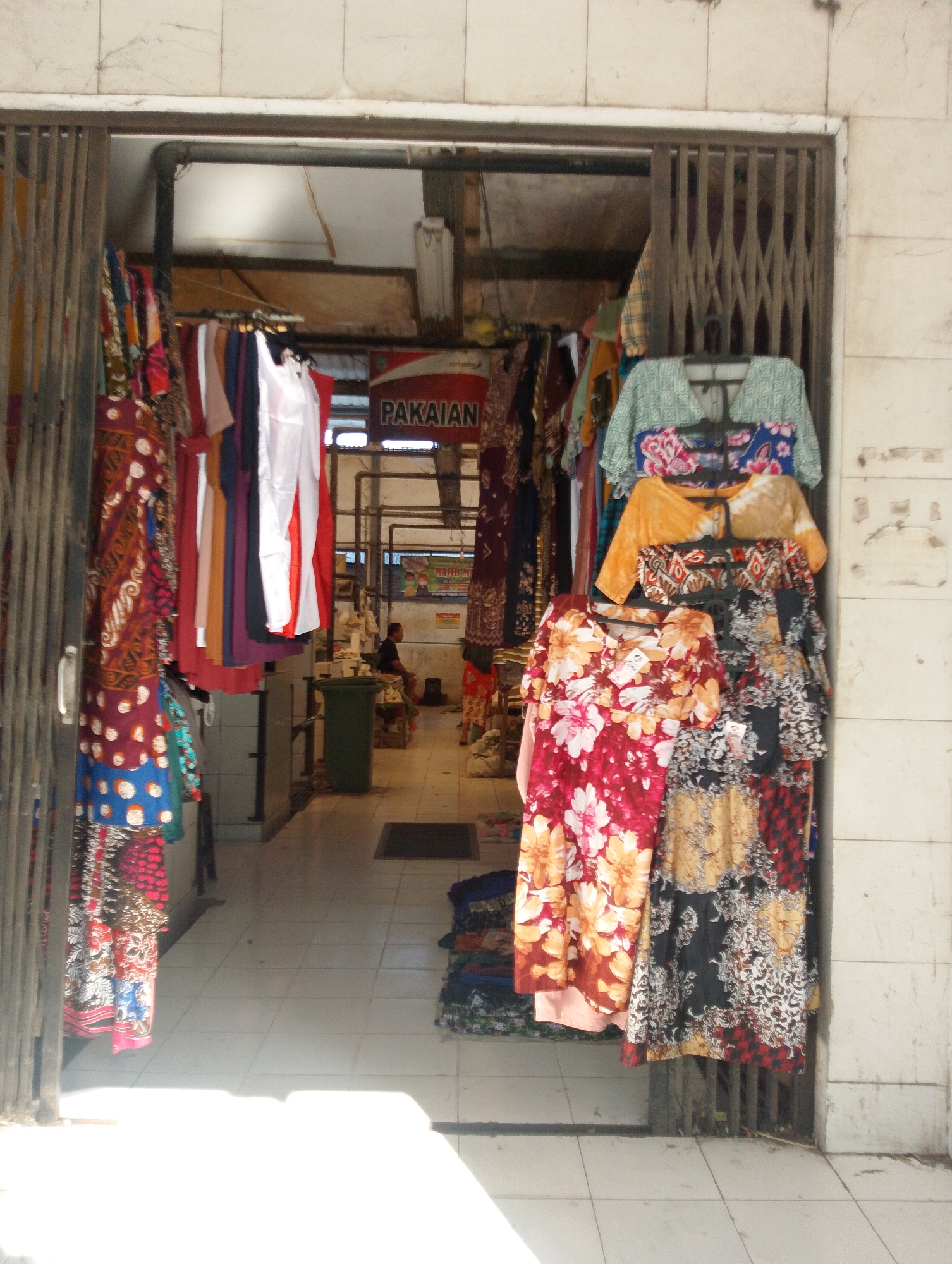 The image shows the entrance to a small clothing store in Kendal Rejo, Indonesia. The store appears to be an outdoor market stall or a small shop with an open entrance. On the left side, there are several racks of brightly colored clothing, including dresses and blouses, with intricate patterns and designs, typical of batik or traditional Indonesian fabrics. The clothing is hung on hangers and displayed outside the shop.
In the background, the interior of the shop is visible, showing more clothing racks and shelves. A sign that reads "PAKAIAN" (meaning clothing in Indonesian) is mounted on the wall inside. Further back, a person is seated, possibly the shopkeeper. The floor is tiled, and the entrance is partially blocked by a metal gate. On the right side of the entrance, more clothing is displayed on hangers, including patterned dresses. The scene suggests a bustling local market environment.