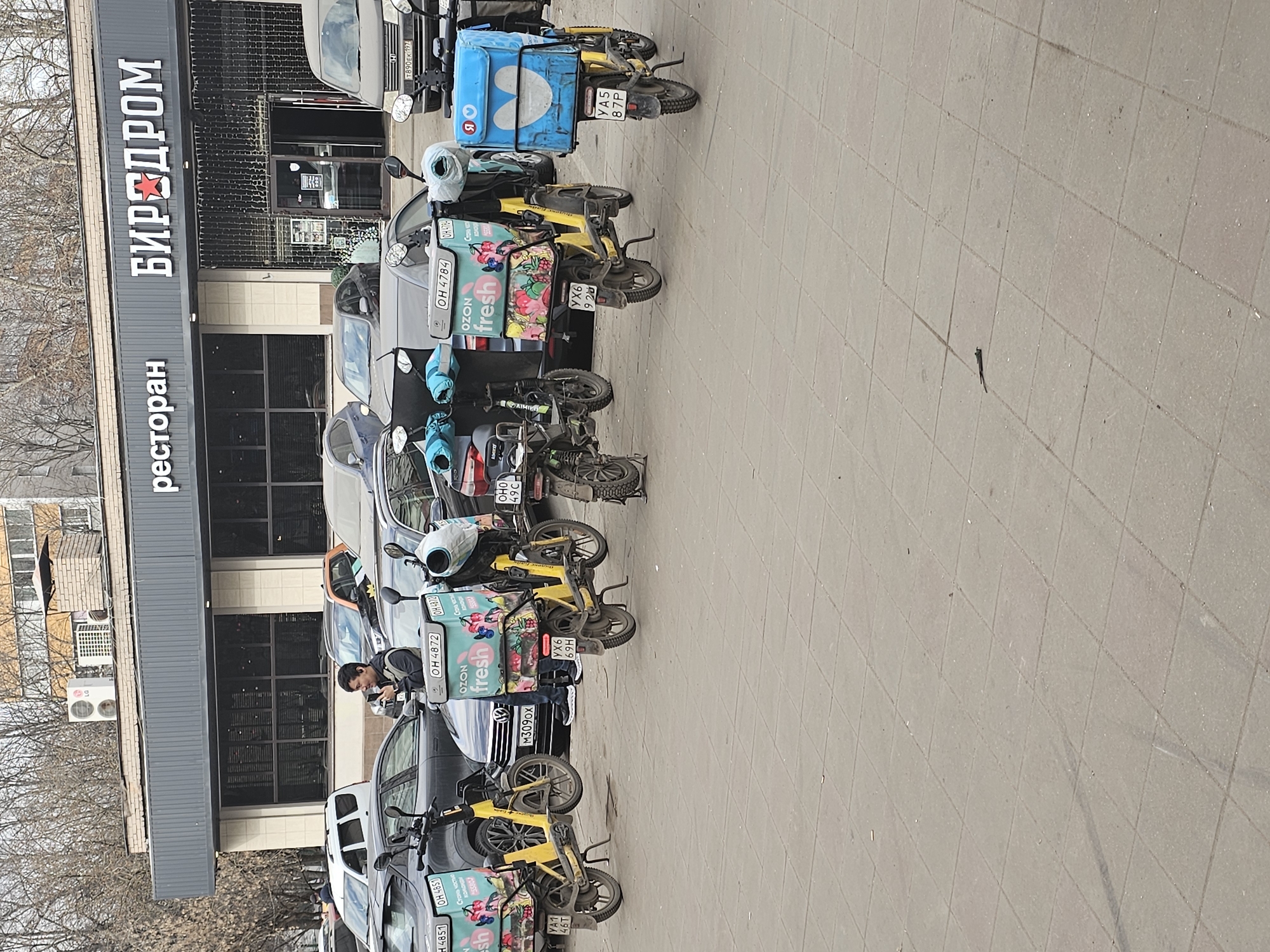 The image depicts a scene outside a restaurant, identified as "БИРДРОМ" (BIRDROME) and translated into English as "Brewery," in Moscow, Russia. Several yellow electric scooters, possibly for delivery, are parked along the sidewalk adjacent to parked cars. The scooters have delivery boxes on their backs, some bearing the "OZON fresh" logo and Russian license plates. A person is seen near one of the scooters. The environment suggests an urban setting with trees visible in the background.