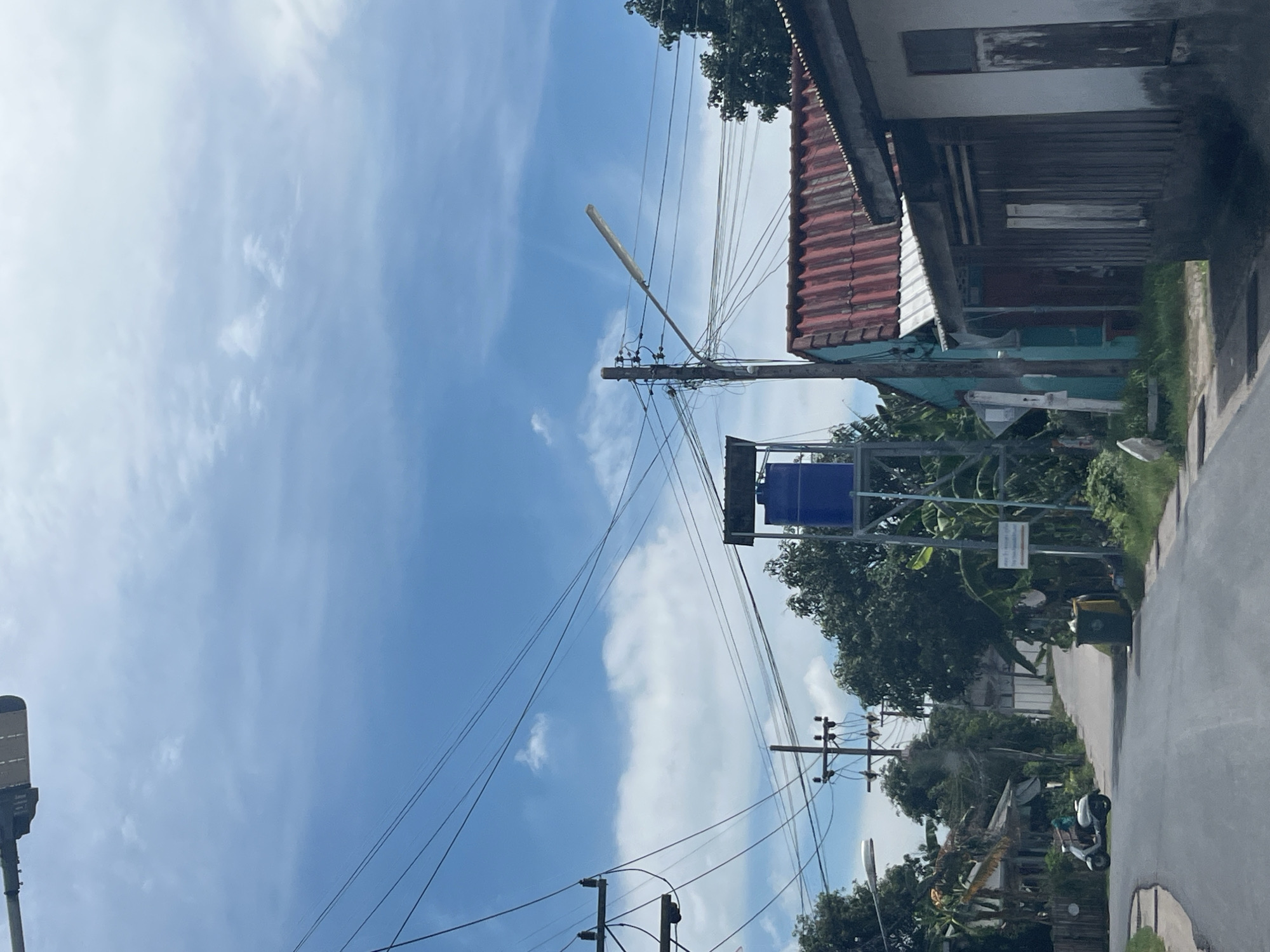 The image depicts a street scene in Rawai, Thailand, under a bright blue sky with scattered white clouds. A paved road curves through a residential area lined with trees and houses. On the left, a white scooter is parked, and a person is partially visible on it. Electrical wires crisscross the sky, supported by utility poles. A tall structure with a blue water tank stands prominently on the right side of the road, near a turquoise building with a red corrugated metal roof. Adjacent to this is a weathered wooden structure. In the distance, more houses and vegetation are visible. Two garbage bins are situated on the side of the road.