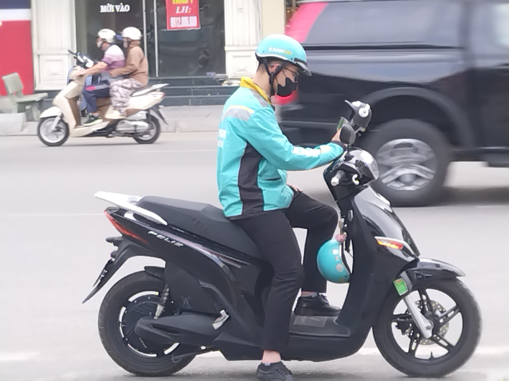 A delivery driver, wearing a distinctive teal and black uniform jacket, black face mask, and a light blue helmet (possibly branded "EAHH"), is paused or moving slowly on a black VinFast Feliz electric scooter. He is looking down towards the handlebars, possibly at a mobile device or controls. A smaller light blue helmet hangs from the left handlebar of his scooter. The scene takes place on a paved urban street, identified as Hong Gai Ward, Vietnam, during daytime under what appears to be an overcast sky.
In the background, two people on a light-colored scooter, both wearing helmets, are moving further down the street. A dark-colored van or SUV is visible, blurred from motion, driving past the main subject. Buildings line the street in the background. One building features a white entrance and a red sign with visible Vietnamese text: "MỜI VÀO" (Welcome/Come in) and "LI: 0812.806.600" (Contact: phone number). The image captures a routine moment of urban life and commerce in Vietnam.