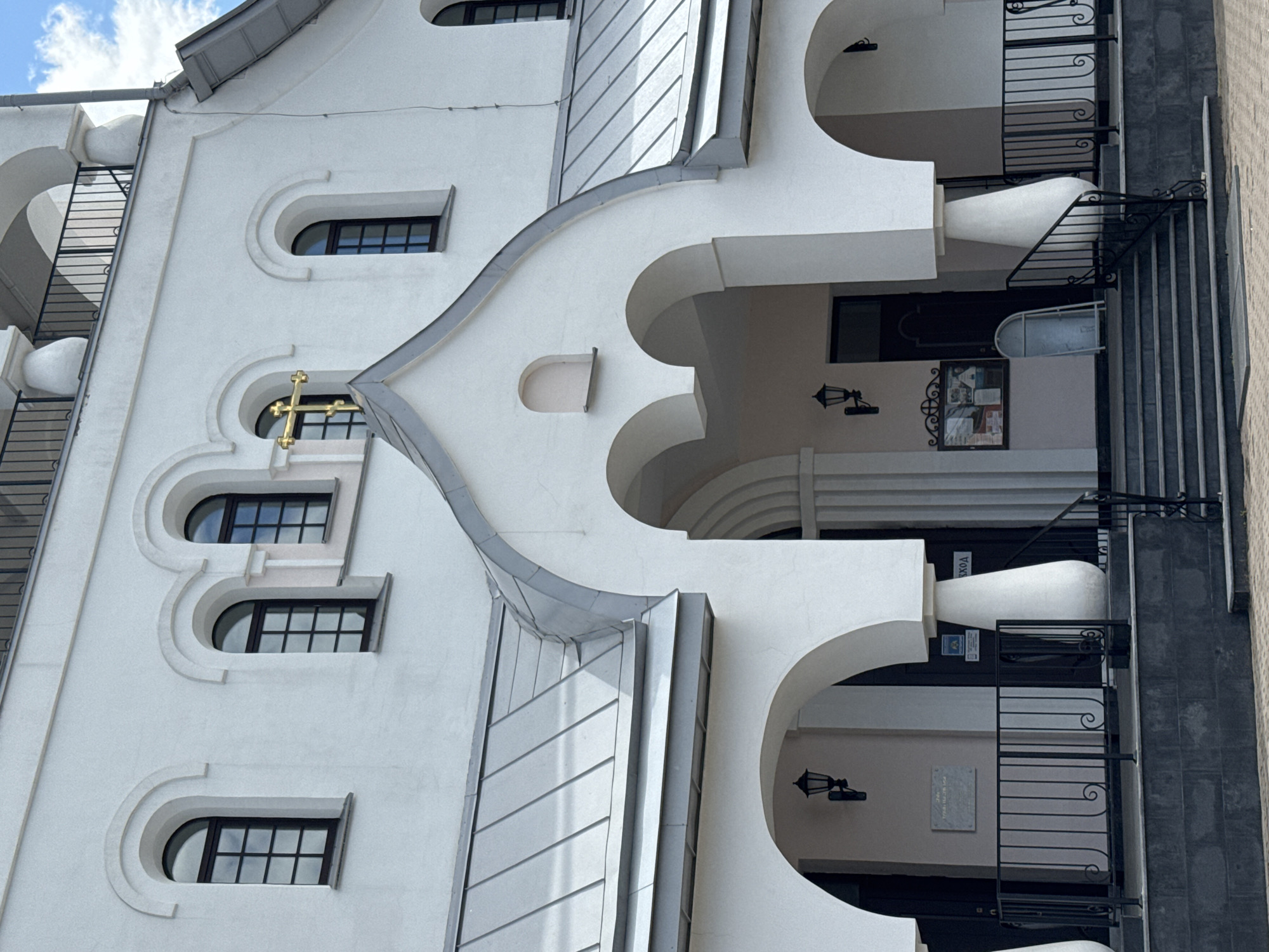 This image shows a white, multi-story building with arched windows and a gray metal roof. A golden cross is visible above one of the windows. The building appears to be a church or religious institution, given the presence of the cross and the architectural style. The entrance features a set of stairs leading up to an arched doorway, with black metal railings on either side. A sign is mounted on the wall near the entrance. The building is set against a backdrop of a partly cloudy blue sky. The context of Saint Petersburg, Russia is not directly evident from the image itself, but the architectural style is consistent with religious buildings found in the city.