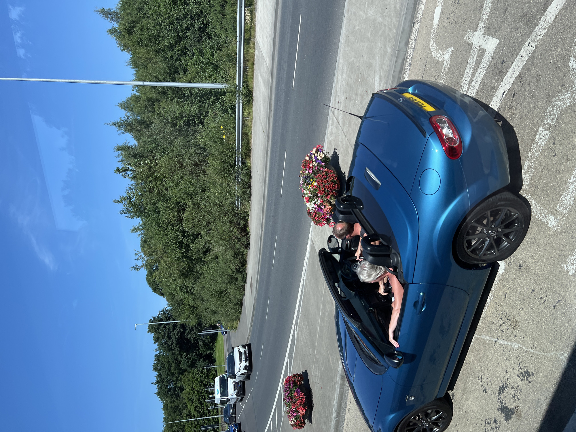 The image captures a sunny day in Letterkenny, Ireland, showing a blue convertible driving on a road. The car, a Mazda MX-5, has its top down and is occupied by two people. A woman with short blonde hair is in the driver's seat, and a man with short brown hair is in the passenger seat. They are both looking forward, suggesting they are traveling.
The car is positioned on the right side of the road, in what appears to be a lane or a turning area. In the background, there's a road with other vehicles, including a white van and a white car. A row of trees and foliage borders the road, and a street light is visible. Flower arrangements are placed on a raised concrete area next to the road, adding a touch of color to the scene. The sky is clear and blue, indicating pleasant weather. The overall impression is a leisurely drive on a bright summer day in Letterkenny.