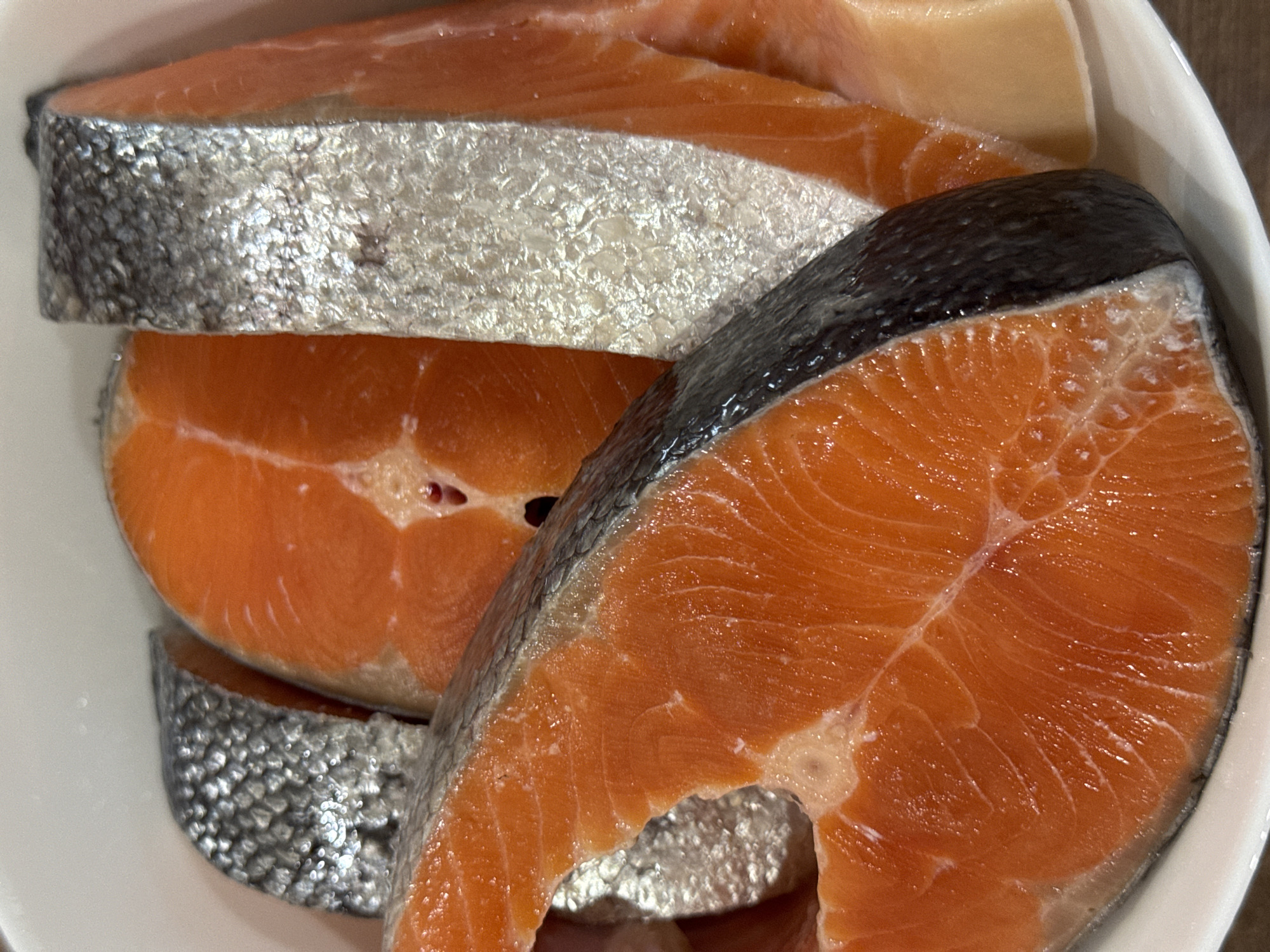 A close-up, overhead shot shows multiple portions of raw salmon steaks arranged in a white bowl. The salmon has a bright orange flesh and is skin-on. Some steaks are cut thicker than others, and the skin is visible as a silvery-grey layer. The bowl is plain white and appears to be ceramic. The lighting is bright, illuminating the glistening texture of the fish. The scene is indoors, likely a kitchen setting, though no other details of the surroundings are visible. The location of Усть-Курдюм, Russia is not determinable from the image.