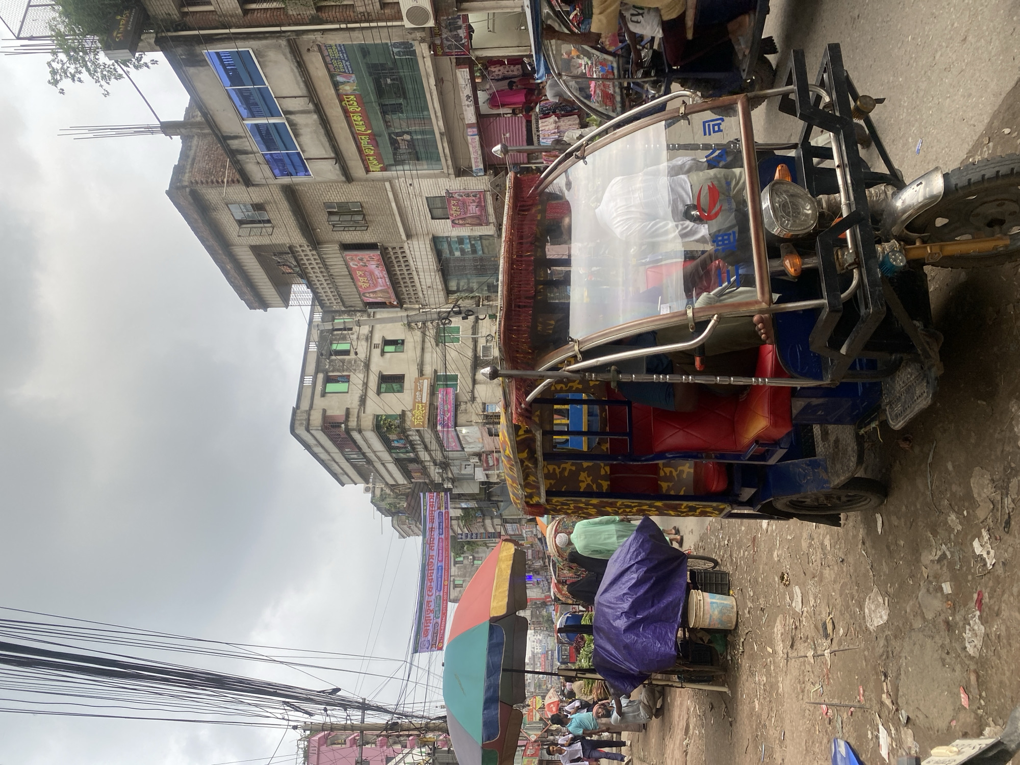 This is a medium shot of a street scene in Dhaka, Bangladesh. The street is crowded with pedestrians and several auto-rickshaws. Buildings line both sides of the street, many of them appearing to be multi-story commercial buildings with shops on the ground floor. Overhead, a tangle of electrical wires crisscrosses the sky, a common sight in Dhaka.
In the foreground, a colorful auto-rickshaw is parked. Its passenger cabin is decorated with a camouflage-like fabric pattern and features bright red seats. The windshield is partially transparent, revealing a person sitting inside wearing a white shirt. The auto-rickshaw's front is equipped with a headlight and a metal rack.
To the left of the auto-rickshaw, a large, colorful umbrella provides shade over a street vendor's stall. A purple tarp covers some goods on a cart. Further down the street, more vendors and shoppers are visible, along with other vehicles, including another auto-rickshaw and what appears to be a bus. The ground is unpaved and littered with debris, indicating a busy and somewhat unkempt urban environment.
Banners and signs with text in Bengali are displayed on the buildings, identifying various businesses and services. The sky is overcast, suggesting a cloudy or possibly rainy day. The overall impression is of a bustling, vibrant, and densely populated urban market area.