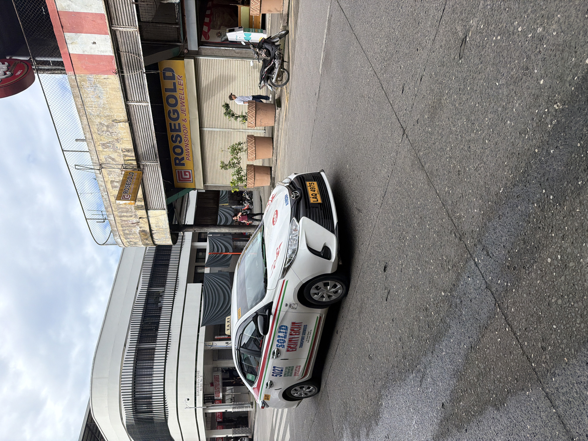 The image captures a street scene in Davao City, Philippines. A white Toyota taxi with plate number LAQ 4975 and the inscription "SOLID EASYRIDE TRANSPORT SERVICES" is parked on the street. On the left side of the frame is the Dakudao & Sons Building. Adjacent to the taxi, storefronts are visible, including one for "ROSEGOLD PAWNSHOP & JEWELLER." A person stands in front of the pawnshop. A motorcycle is parked nearby. The street appears wet. The sky is overcast.