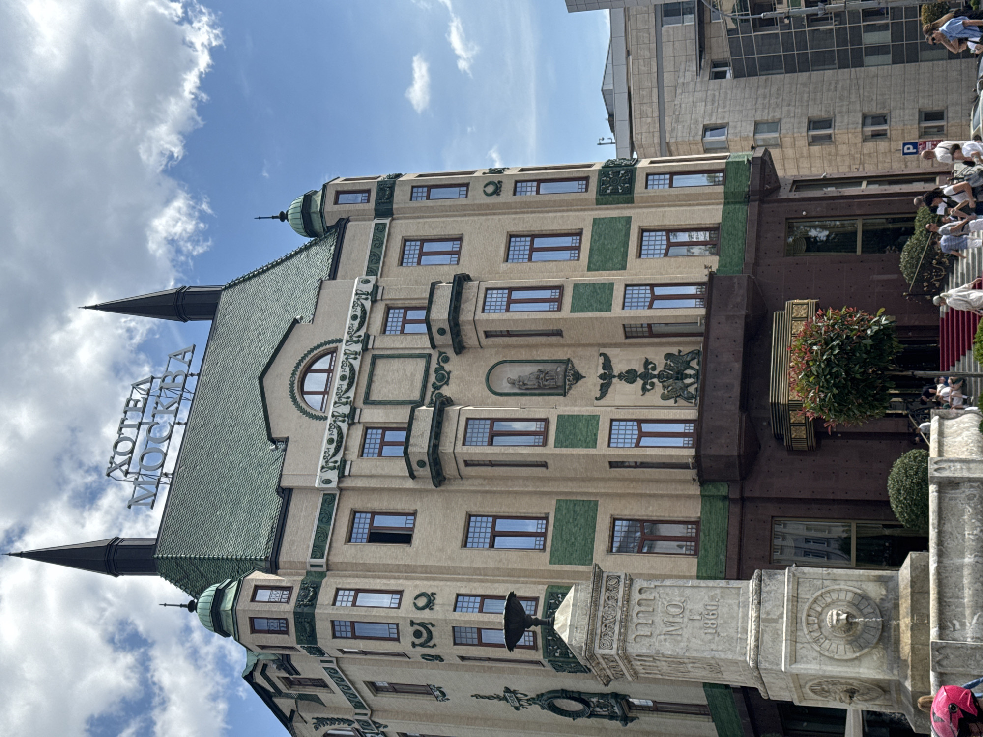 The image shows the Hotel Moskva in Belgrade, Serbia, a historic landmark building with distinctive architecture. The hotel's facade is light beige brick with green decorative elements and a green tiled roof. Two black spires top the building. A sign reading "HOTEL MOSKVA" in Cyrillic letters is visible on the roof. In the foreground, a stone monument with a lion's head fountain is present, along with a red carpet leading up to the hotel entrance. Several people are visible, some walking on the sidewalk and others gathered near the entrance. The sky is blue with scattered white clouds. The surrounding area includes other modern buildings, suggesting an urban setting.