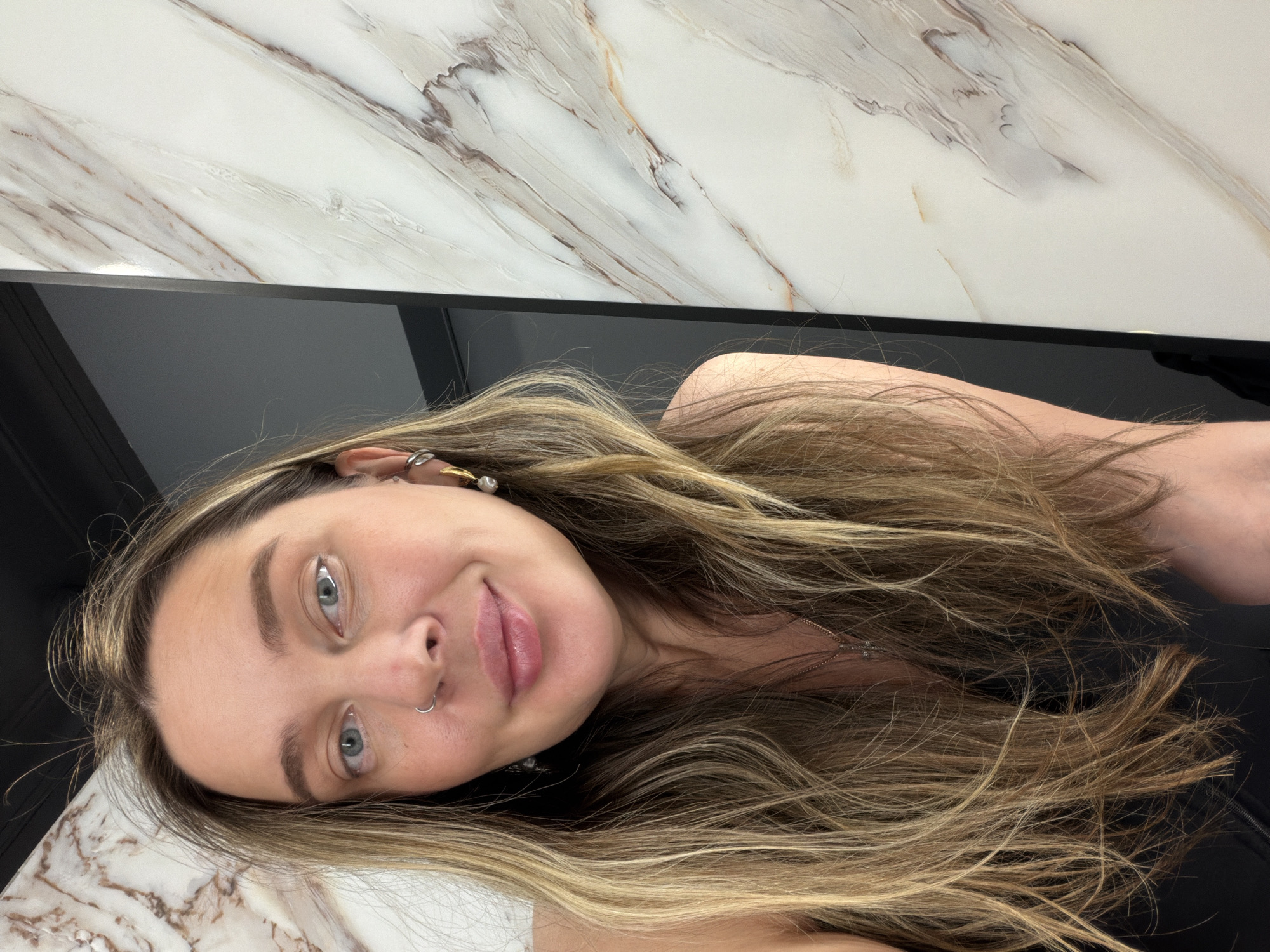 A fair-skinned woman with long, highlighted blonde hair is pictured in a close-up shot. She has light blue eyes and a nose ring. Her expression is playful, with a slight smirk and her tongue gently touching her upper lip. She is wearing earrings. Her attire appears to be black. The background consists of a white and gray marble-patterned surface above a black surface. The picture is taken at a tilted angle. The scene does not contain any context that would indicate Moscow, Russia.