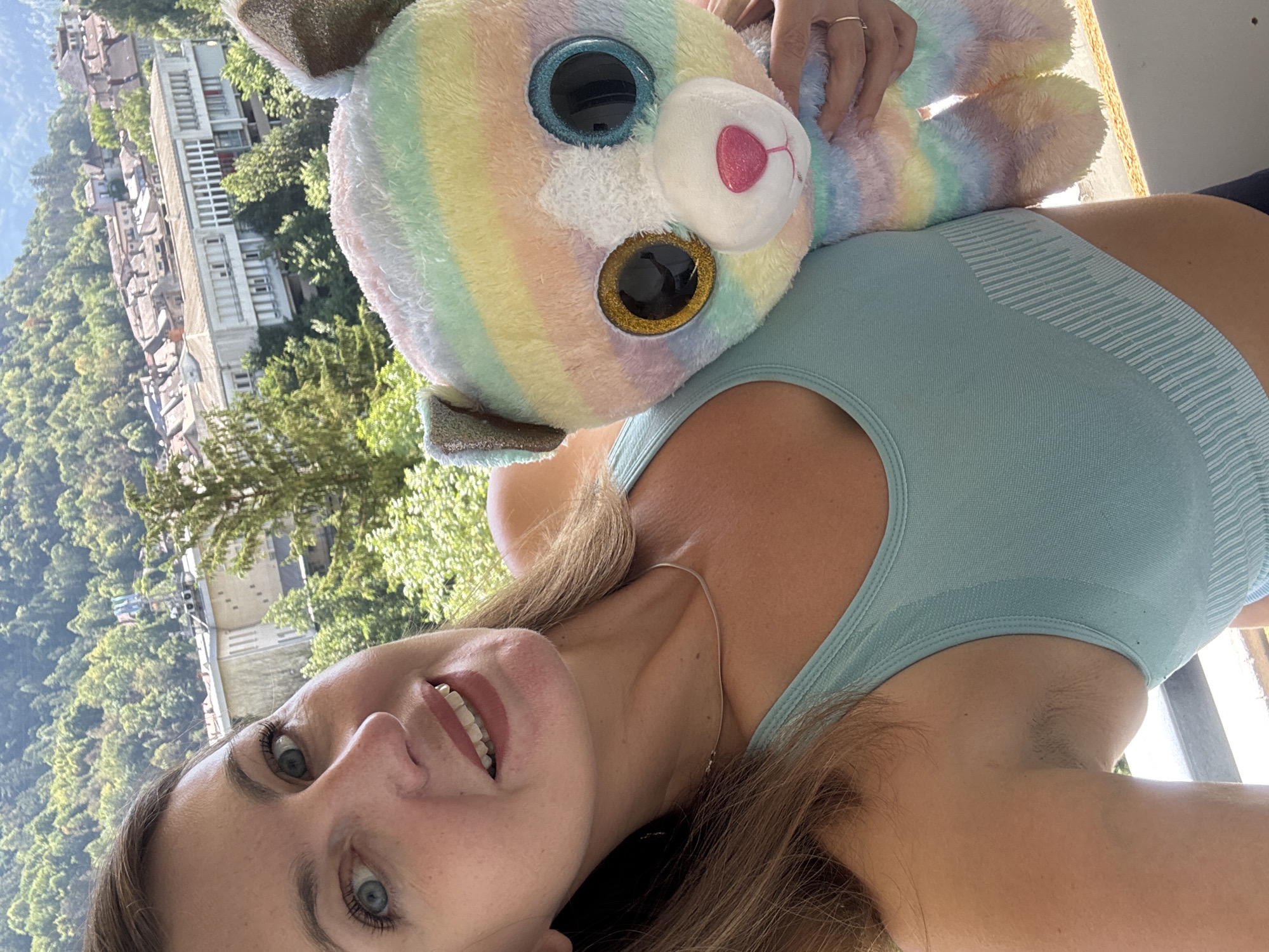 A woman with light brown hair and blue eyes takes a selfie while holding a colorful, rainbow-striped plush toy. She is wearing a light blue sports bra and a thin silver necklace. Her left arm is bent, with her hand resting on the plush toy's back. The background shows a town with buildings and trees, illuminated by sunlight. The perspective suggests the scene is on a balcony or elevated area. The town's architecture, with its tiled roofs and multi-story buildings, is characteristic of Saint-Claude, France. The surrounding hills are covered in lush green trees.
