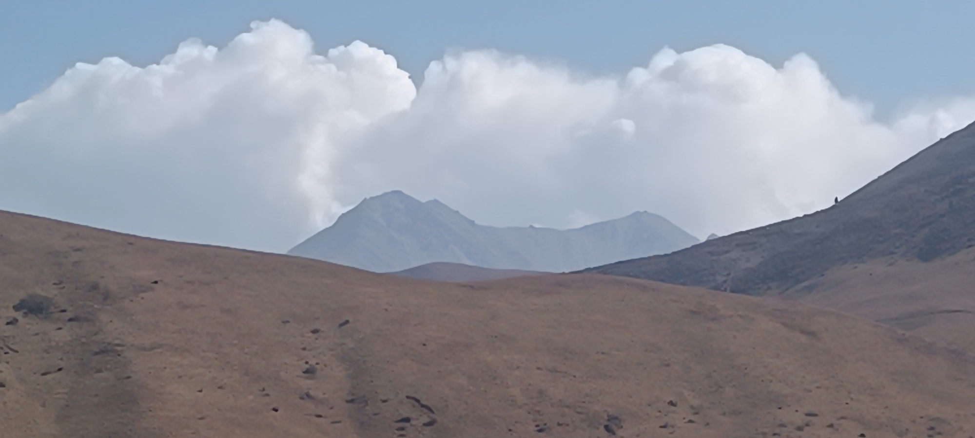 An outdoor, medium-shot photograph captures a distant view of a mountain range under a partly cloudy sky. The foreground consists of a reddish-brown, barren mountain slope dotted with small rocks and shadows. In the mid-ground, rolling hills of the same color transition into the darker, rougher terrain of another mountain slope. On the far right side of the image, a lone, small, dark silhouette of a person is visible walking up this slope. In the background, a jagged, blue-gray mountain peak rises, partially obscured by large, fluffy white clouds. The sky is a pale blue, with the sun's light filtering through the clouds. The overall scene evokes a sense of vastness and solitude in a mountainous landscape.
