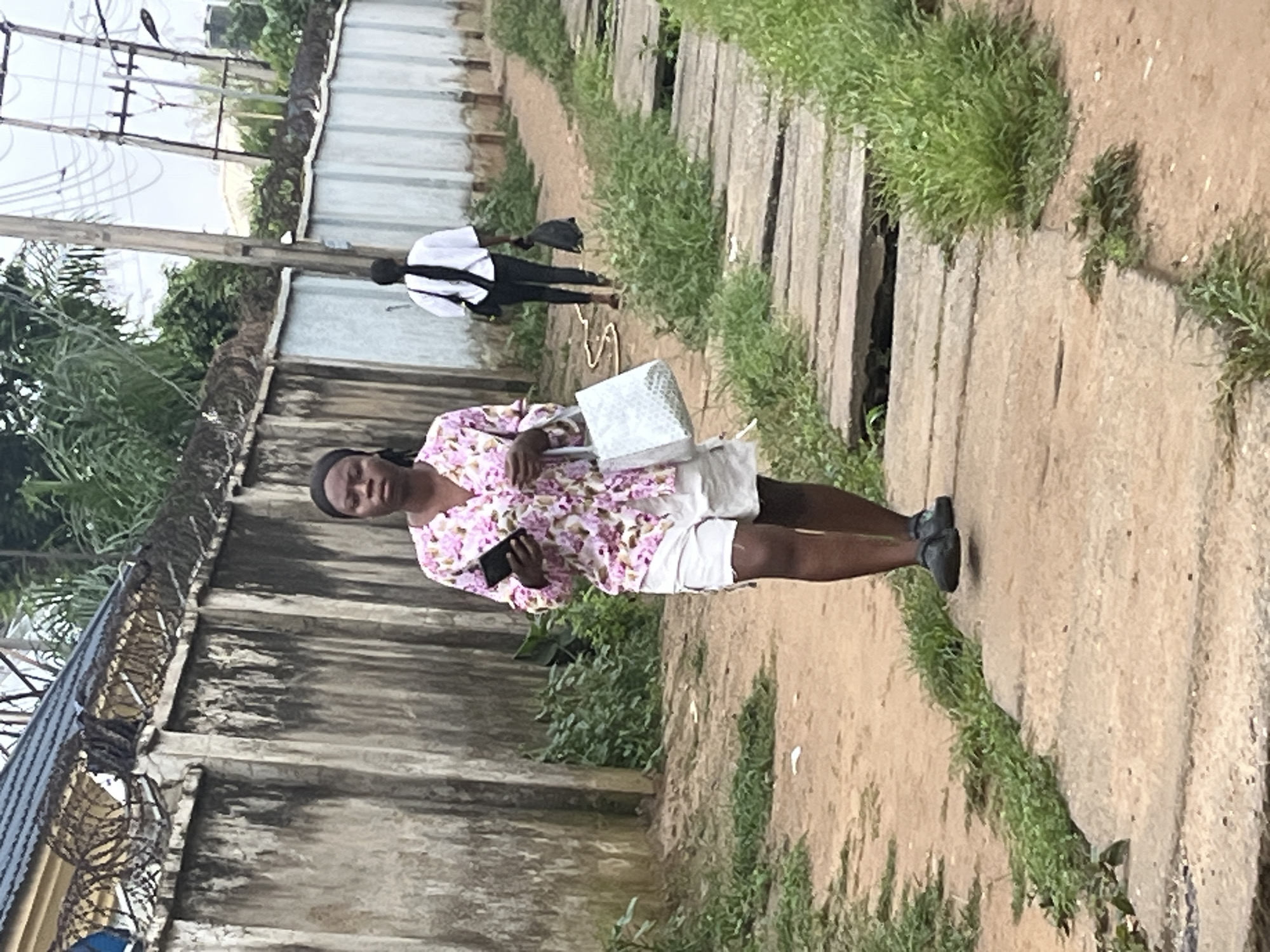 An African woman walks on a concrete path next to a dirt road, holding a phone and a silver bag. She wears a pink floral shirt and white shorts. Behind her, another woman in a white shirt and black pants walks away, carrying a black bag. The scene is in Abuja, Nigeria, with a concrete wall and barbed wire fence on the left, and lush vegetation and utility poles in the background.