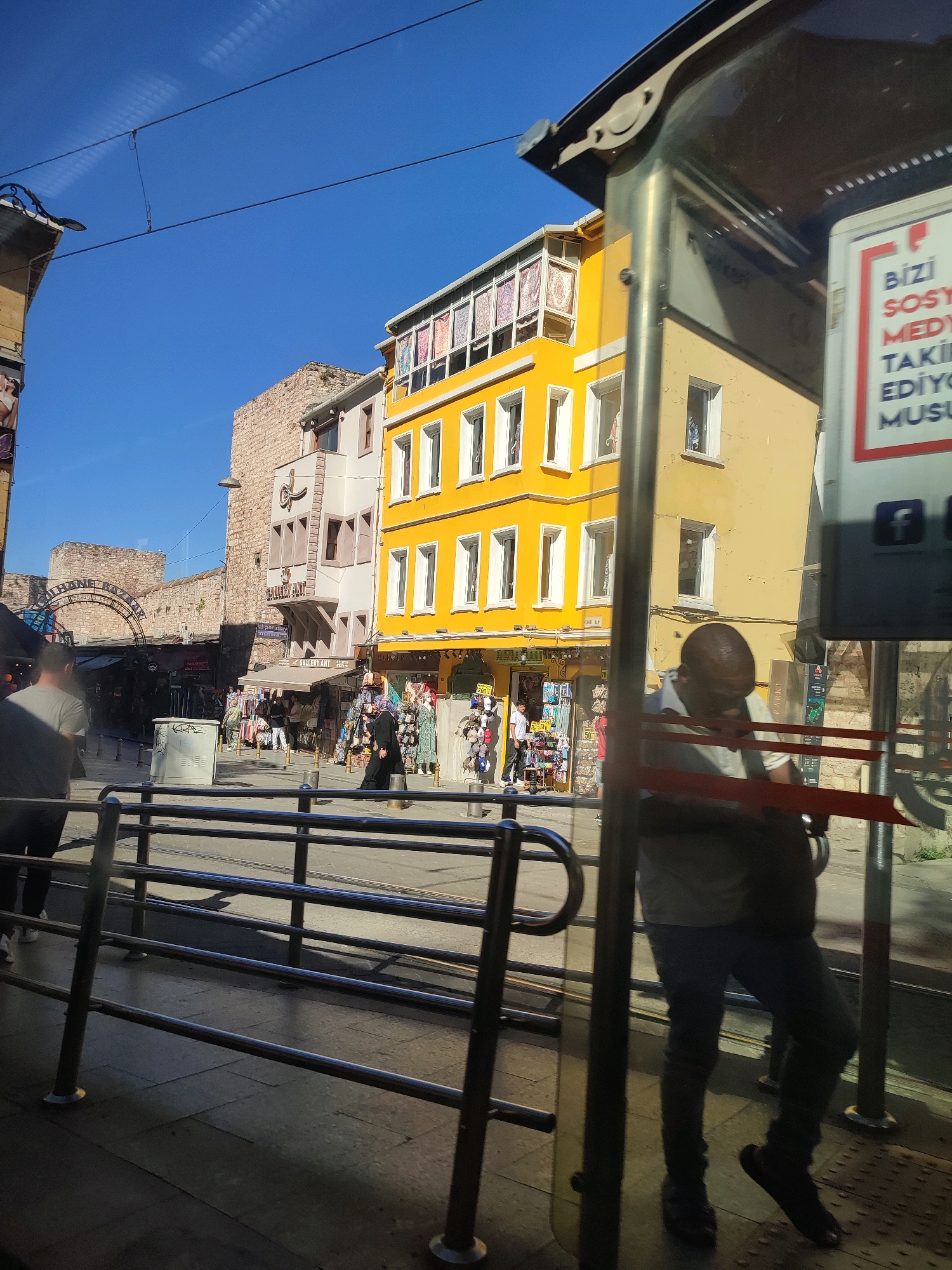 The image depicts a street scene in Istanbul, Turkey, likely near the Grand Bazaar or a similar market area, given the architecture and the presence of shops. In the foreground, a metal railing system, possibly part of a public transit stop or pedestrian walkway, is prominent. A person is visible behind the railing, looking down and holding something in their hands. The background features brightly colored buildings, a yellow one being particularly noticeable with multiple windows. Shops are visible at street level, displaying goods and signage. People are walking in the middle ground, and in the distance, more traditional brick architecture is evident. The sky is clear and blue. The overall impression is a busy urban environment with a blend of historical and modern elements.