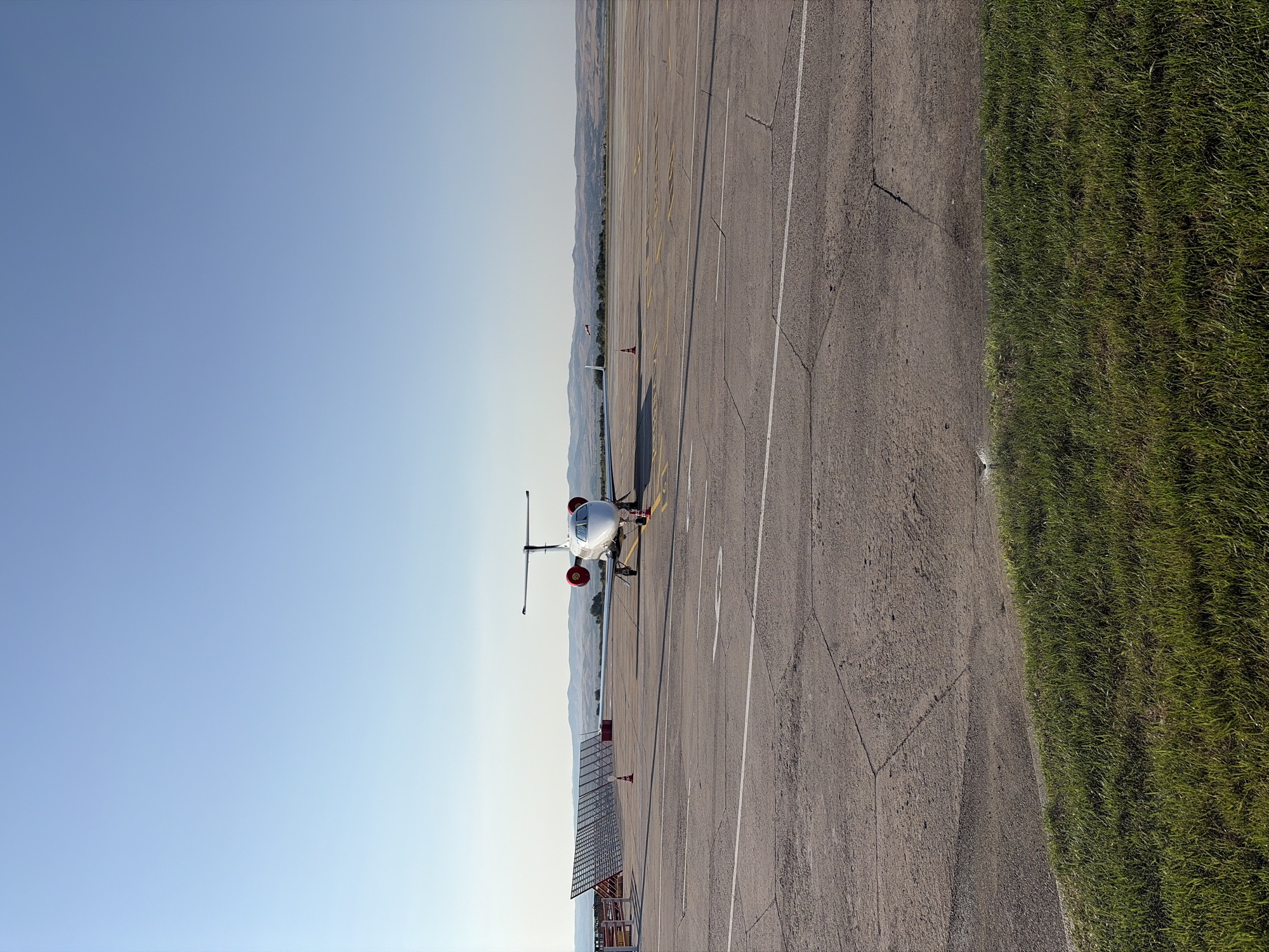 A corporate jet airplane is parked on a tarmac at an airport under a clear blue sky. The airplane is white with red engine nacelles and has its nose facing the camera. Orange cones are placed around the aircraft. In the background, there are distant hills and vegetation. In the left midground, there's a metal structure with a grid pattern, possibly part of an airport fence or equipment. The foreground consists of cracked asphalt and a strip of green grass. The image appears to be taken on a sunny day. This scene is at Burgas Airport, Burgas, Bulgaria.