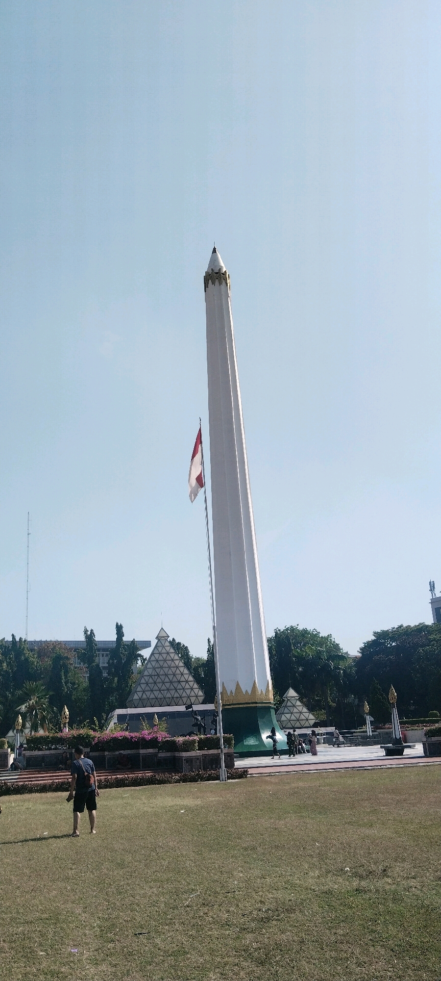 The image depicts the Tugu Pahlawan monument in Surabaya, Indonesia, under a clear blue sky. The monument, a tall, white, obelisk-shaped structure with gold accents at the base and apex, dominates the scene. An Indonesian flag, red and white, flies from a flagpole near the monument.
In the foreground, a grassy field has scattered patches of dry grass. A person wearing a dark t-shirt and shorts is walking away from the camera towards the monument. Further back, near the monument's base, a group of people are gathered. One person is wearing a light-colored dress, and others are in casual attire.
Behind the monument and to its left is a distinctive pyramid-shaped building with a geometric facade, also a part of the Tugu Pahlawan complex. Green trees line the background, with some buildings visible beyond them. The environment appears to be a public park or plaza, designed for commemoration and recreation. The lighting suggests it is daytime, likely late morning or early afternoon, given the angle of the sun and the bright conditions.