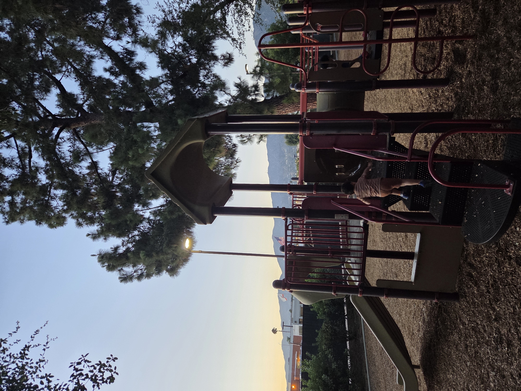 At dusk, a young boy with brown shorts and a striped shirt climbs the stairs of a playground in Glendale, California. The playground features a slide, tunnels, and red metal railings. A tall pine tree with a lit lamp post beside it dominates the background. In the distance, mountains are visible under a clear, pale sky, with the American flag flying in the background. The ground of the playground is covered in wood chips.