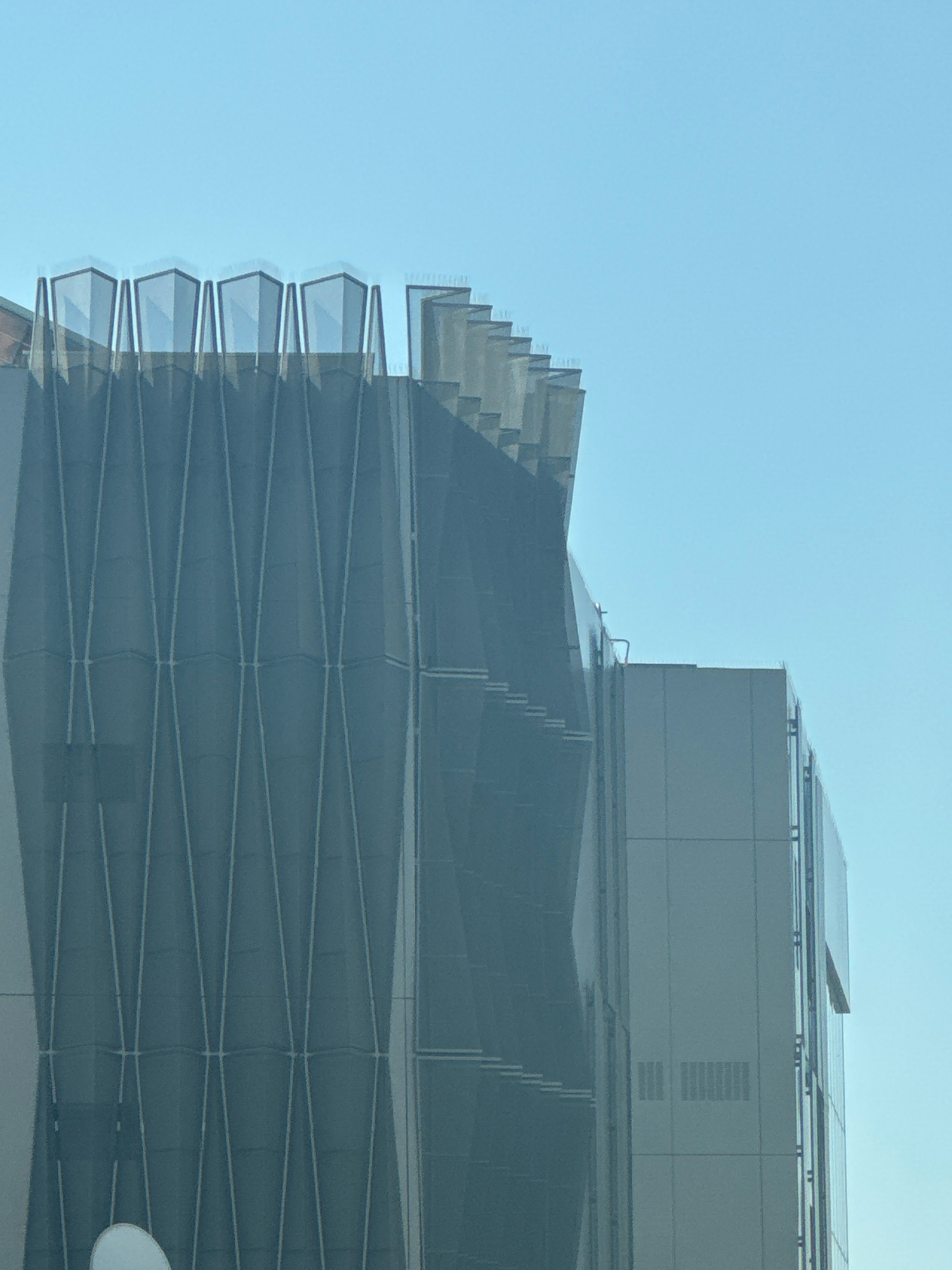 The image captures the upper section of a modern building with a distinctive architectural design, set against a clear blue sky. The building's facade is characterized by a dark, textured material with a geometric pattern of intersecting lines, forming a diamond-like grid. Above this patterned section, the building's roofline features a series of angular, translucent elements, possibly made of glass or a similar material, that project upwards and outward in a staggered arrangement. To the right of this main structure, another section of the building is visible. This part has a smoother, lighter colored facade composed of large rectangular panels. The sky is cloudless and a uniform pale blue, indicating a bright day. The image appears to be taken from a low angle, looking up at the building. There are no people or active elements visible in the frame, giving the scene a static and architectural focus. The specific location is the city of Mesa Geitonia, Cyprus.