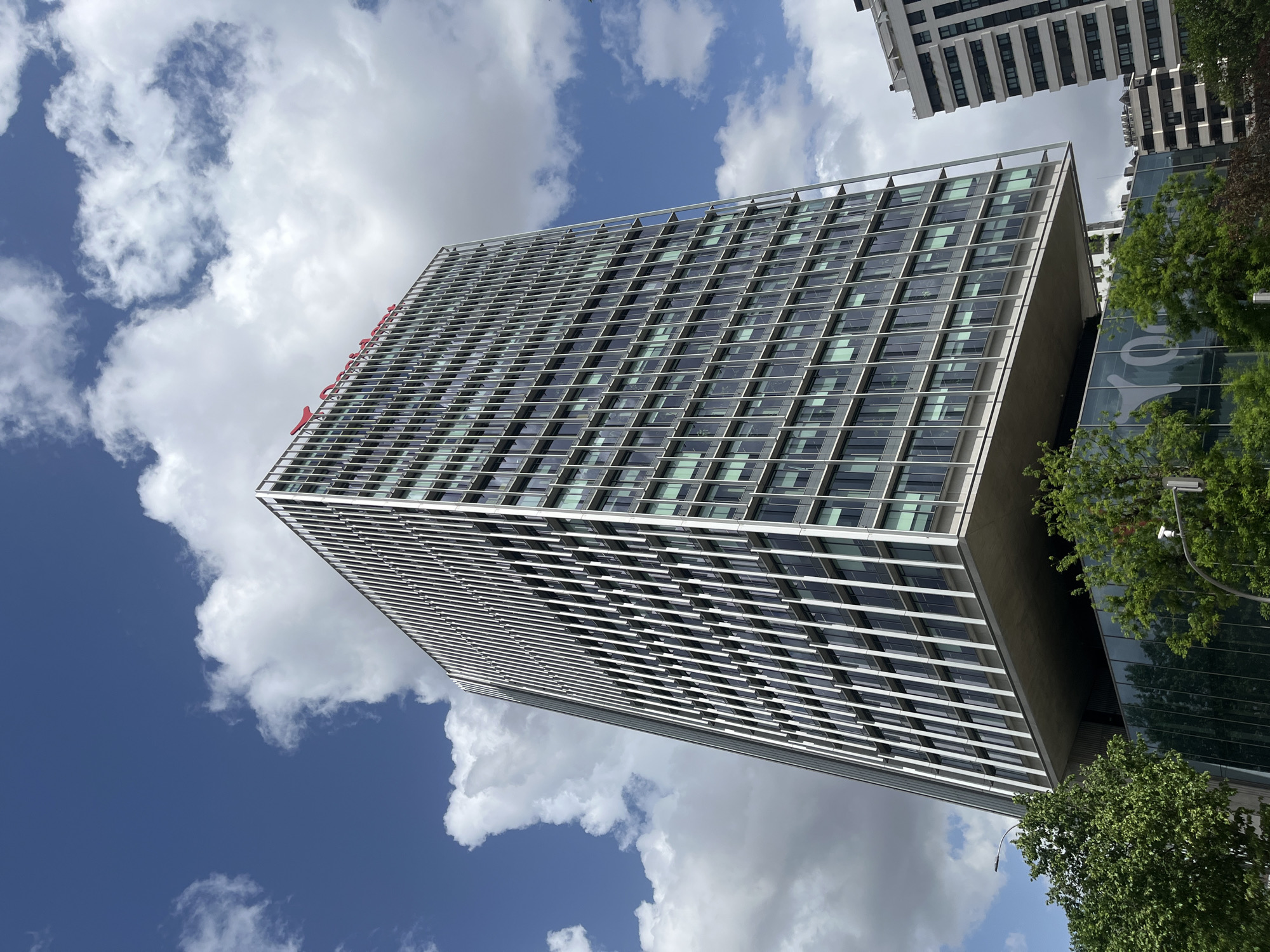 The image displays a modern, tall building set against a bright blue sky dotted with scattered white clouds, captured from a low-angle perspective. The main building features a sleek, rectangular design with a facade characterized by a grid pattern of dark-tinted windows and numerous horizontal, light-colored architectural elements that may function as sunshades.
On the upper portion of this prominent building, red lettering is clearly visible, spelling out "Meliá". To the right of the main structure, another contemporary building with a glass facade is partially visible. This secondary building features large, light-colored or white text, which is partially obscured but appears to contain characters such as "00" or "O0" and potentially parts of a word like "Galicia" due to visible letter fragments. Further behind and to the right, other multi-story buildings with balconies can be seen.
Green trees with dense foliage are present in the foreground and near the base of the buildings, providing a natural contrast to the urban architecture. The bright daylight and clear sky indicate a daytime setting, likely late morning or early afternoon. No individuals are visible in the scene. The overall impression is of a modern urban environment in a city like Madrid, Spain.