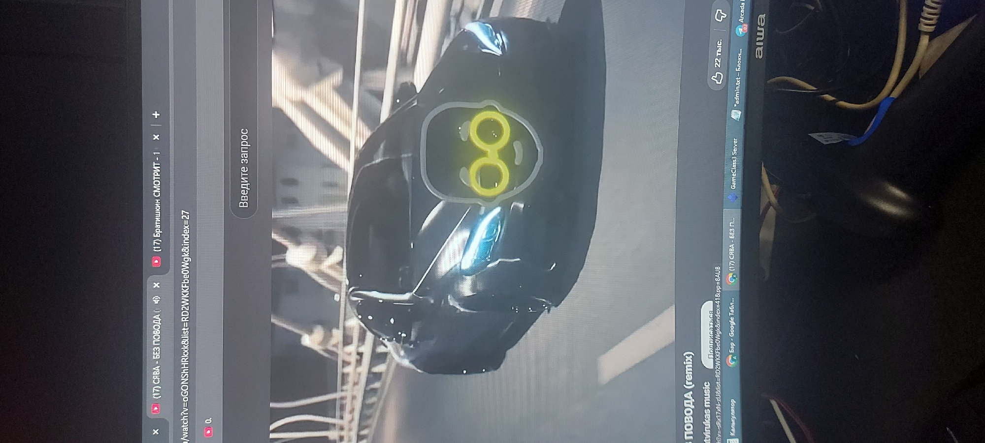 The image shows a computer monitor displaying a partially visible video of a black object with glowing yellow circles on it. Tabs at the top of the monitor show web pages, including YouTube videos with titles in Russian: "(17) СЯВА - БЕЗ ПОВОДА" and "(17) Братишкин СМОТРИТ - 1". Other open applications are visible on the right side of the screen, including "GameClass3 Server" and "*admin.txt - Блокнот...". The monitor is surrounded by cables and the edge of a desk or surface. The location is not discernible from the image content.