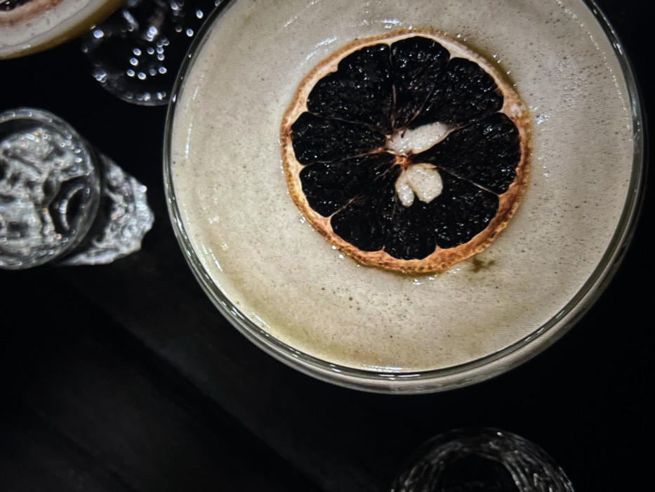 A cocktail, likely a sour, is presented in a coupe glass. A dehydrated citrus wheel, darkened due to dehydration, adorns the foamy surface. A partial slice of citrus fruit is positioned at the top-left edge of the frame. Two etched glasses are on a dark surface and appear to hold water. The scene is dimly lit, highlighting the cocktail and accentuating the dark backdrop.