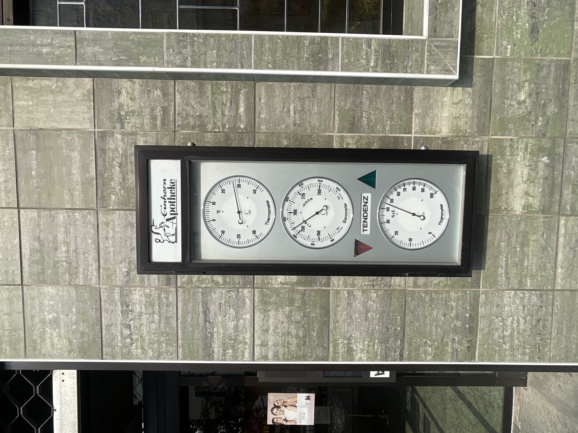 The image shows an outdoor meteorological instrument mounted on a tiled wall. The instrument is housed in a dark frame and displays three gauges: a thermometer, a barometer, and a hygrometer. The thermometer is at the top, measuring temperature in degrees Celsius. Below it is the barometer, indicating atmospheric pressure in Torr. The hygrometer, at the bottom, measures relative humidity in percent (% r.F.). The instrument has a label that reads "Einhorn Apotheke," identifying it as belonging to the Einhorn Pharmacy. The wall is covered with square, light green tiles with a textured, weathered appearance. To the left of the instrument, a glass door with a metal grate is visible, and a poster featuring three women is affixed to the door. To the right, a window with metal bars is present. The scene suggests an urban setting, specifically in Iserlohn, Germany, as indicated by the "Einhorn Apotheke" sign. The lighting suggests it is daytime, with sunlight casting shadows and highlighting the textures of the wall. No people are actively engaged in any activity within the frame.