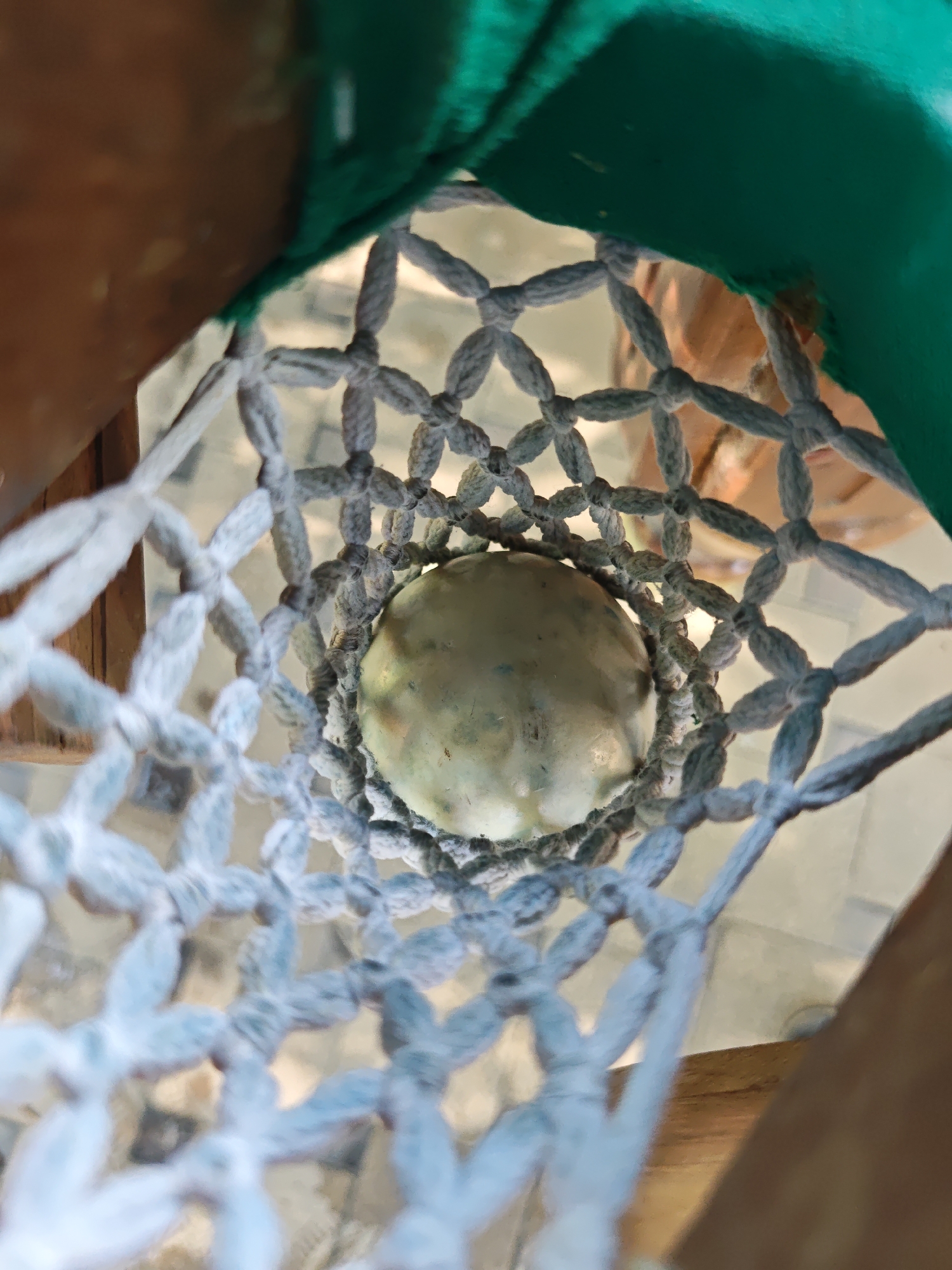 The image is a top-down, close-up shot of a single, light-colored, spherical object, possibly a fruit or vegetable, suspended within a macrame net. The net is made of thick, light-colored cords, forming a diamond pattern. The object is roughly centered within the net. Beyond the net, blurred background elements suggest an outdoor setting, with hints of wooden structures and possibly reflective surfaces. A bright green fabric is visible at the top of the frame, partially obscuring the view. The lighting appears to be natural daylight. The image is taken from a low angle, looking upwards through the net towards the object. The context of Yakkatut, Uzbekistan, is not directly discernible from the visual information within the image itself.