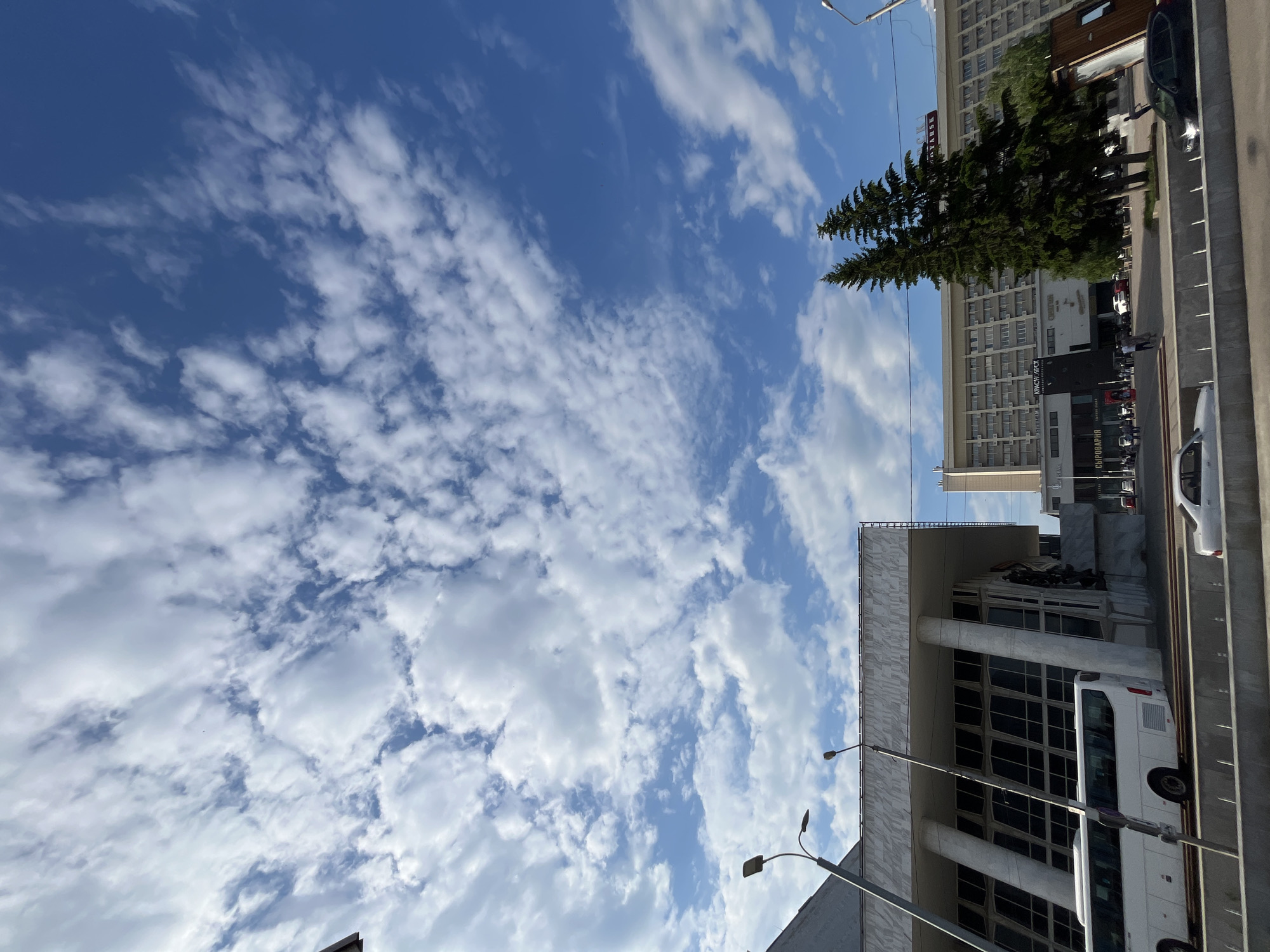 This image captures a daytime exterior view of a city street in Krasnoyarsk, Russia, characterized by a bright blue sky filled with scattered white clouds. In the foreground, a gray concrete barrier lines a street, with a white bus and a white sedan visible on the road. Behind the barrier, a modernist building with a facade of white stone and large, angled windows stands to the left. A tall street lamp is positioned in front of this building. To the right of this structure, a larger, multi-story hotel or office building with a patterned facade dominates the middle ground, flanked by two tall evergreen trees. The hotel features signage, including one that reads "KRASNOYARSK" in large letters at the top. A dark-colored car is parked in front of the hotel. People are walking on the sidewalk in front of the hotel. The overall scene depicts an urban environment with a mix of architectural styles under a partly cloudy sky.