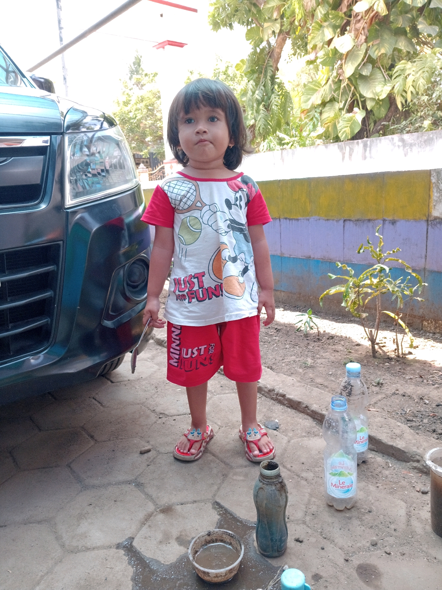 A young child stands in front of a dark-colored SUV in Gebang, Indonesia. The child is wearing a red and white t-shirt with a cartoon character and red shorts with "JUST FOR FUN" printed on them, and red flip-flops. The child is holding a metal spoon in their right hand. Several plastic water bottles and a small bowl of liquid are on the ground near the child. The background includes a colorful wall, trees, and a dirt ground. The scene appears to be an outdoor residential area.