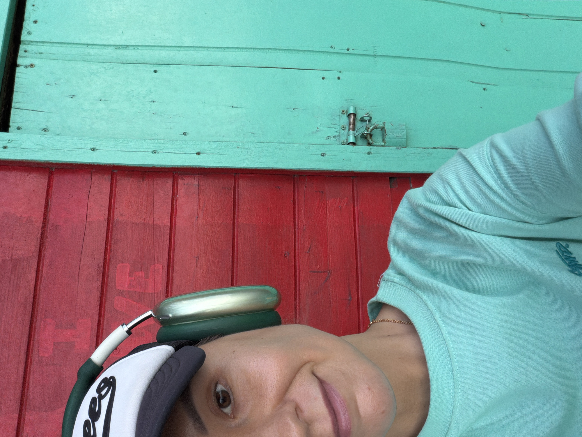 A close-up, selfie-style, waist-up shot features a person wearing a teal sweatshirt, a black and white baseball cap, and green headphones. The background is divided into two distinct sections: a vibrant red wooden wall on the left, with faint, large white lettering that appears to spell "LOVE" horizontally, and a bright teal-colored wooden door on the right. The door has visible hinges, screws, and a padlock securing it. The person is looking towards the camera with a slight smile. The lighting suggests it is daytime, with the sun casting a bright light on the scene. The image appears to have been taken outdoors. There are no clear indicators to place the location specifically within Nha Trang Ward, Vietnam, but the aesthetic suggests a potentially tropical or coastal environment given the bright colors.