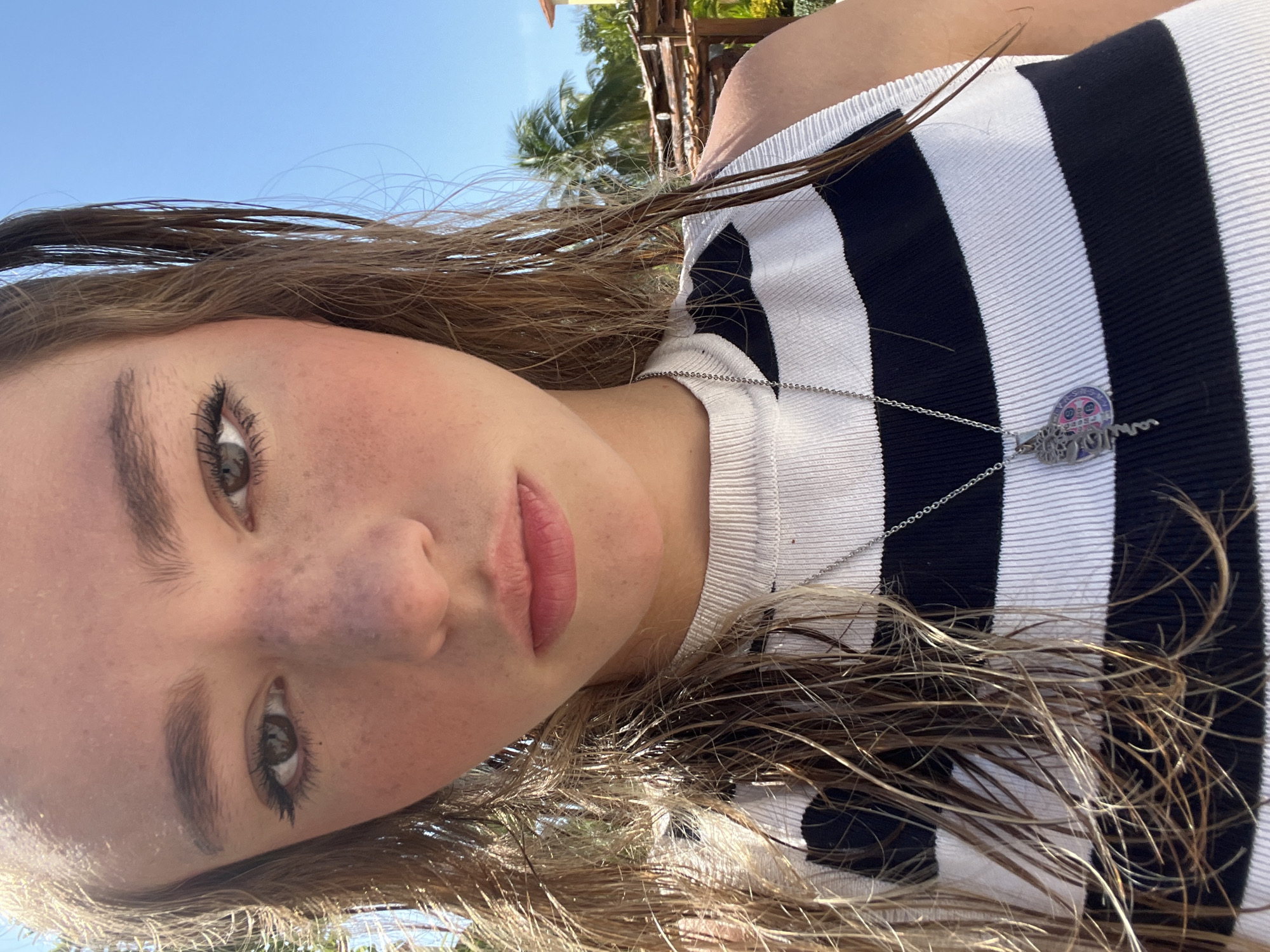A young woman with light brown hair and grey eyes poses in an outdoor location with palm trees and a wooden structure visible in the background. She wears a black and white striped top and a silver necklace with a decorative pendant. Her hair appears to be wet and unkempt. The lighting suggests it is daytime. The location is Playa del Carmen, Mexico, based on the visual context of palm trees.