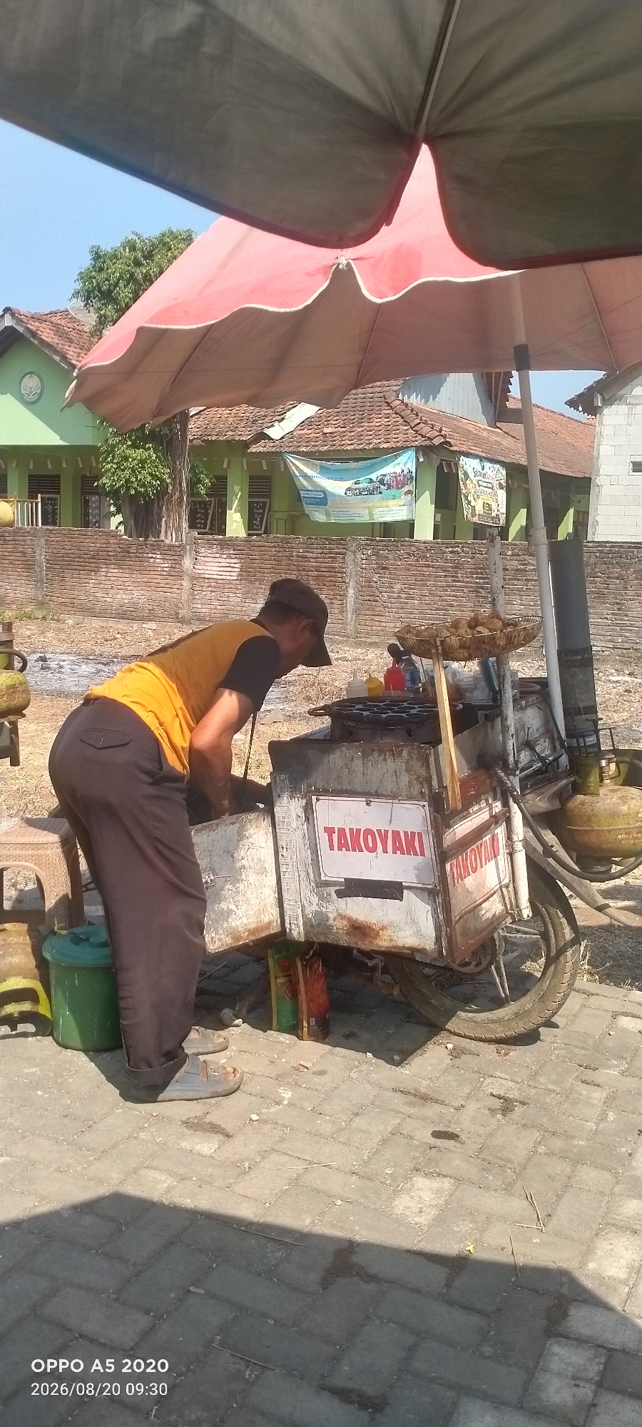 The image depicts a street scene in Sowan Kidul, Indonesia, under a large umbrella. A man wearing a brown cap and an orange and black t-shirt is bent over a food cart that appears to be selling takoyaki, indicated by a sign on the side of the cart. The cart is equipped with a cooking surface with multiple circular molds, a basket of food, and two gas cylinders. Beside the cart, there is a green bucket and a small stool. In the background, a building with a green facade and a tiled roof is visible, along with a brick wall and trees. The ground is paved with interlocking concrete blocks. The overall impression is of an outdoor food vending setup on a sunny day.