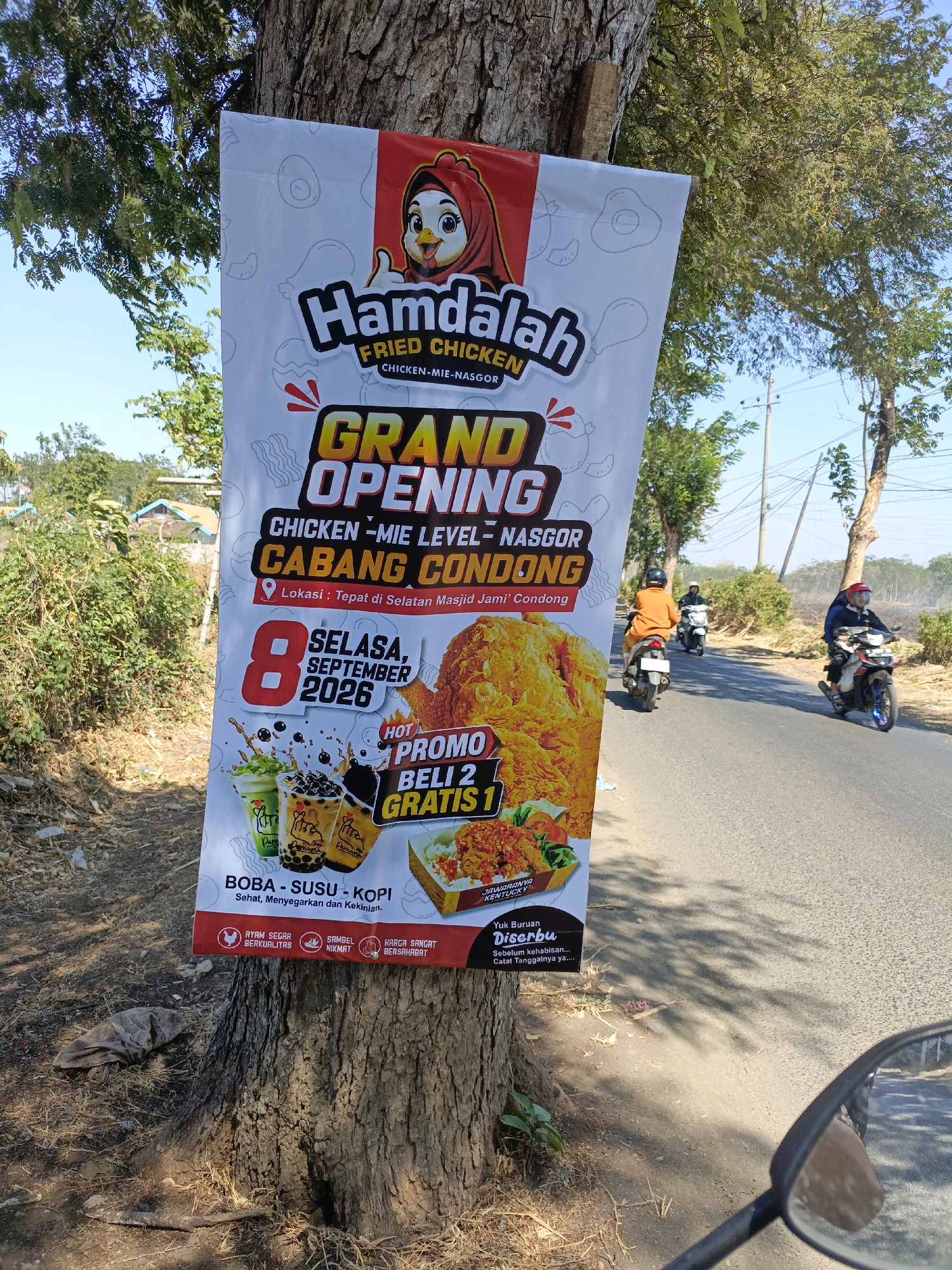 An outdoor scene depicts a large banner for "Hamdalah Fried Chicken" attached to a tree by the side of a paved road. The banner is primarily white with red and yellow accents and features graphics of fried chicken, drinks, and text. Key text on the banner includes "GRAND OPENING," "CHICKEN - MIE LEVEL - NASGOR CABANG CONDONG," a location pin icon with "Lokasi: Tepat di Selatan Masjid Jami' Condong," and a date "8 SEPTEMBER 2026." There is also a "HOT PROMO BELI 2 GRATIS 1" offer and images of three drinks, likely boba, milk, and coffee. The bottom of the banner has icons and text related to "AYAM SEGAR BERKUALITAS," "SAMBEL NIKMAT," and "HARGA SANGAT BERSAHABAT." A small section of text at the bottom reads "Yuk Buruan Diserbu. Sebelum kehabisan... Catat Tanggalnya ya....". In the background, there are trees, some sparse vegetation, and a few small buildings. Several people on motorcycles are visible on the road, some of whom are moving away from the camera. The overall environment appears to be a rural or semi-rural setting in Indonesia, near Condong. The lighting suggests it is daytime with clear skies. A motorcycle's side mirror is visible in the foreground.