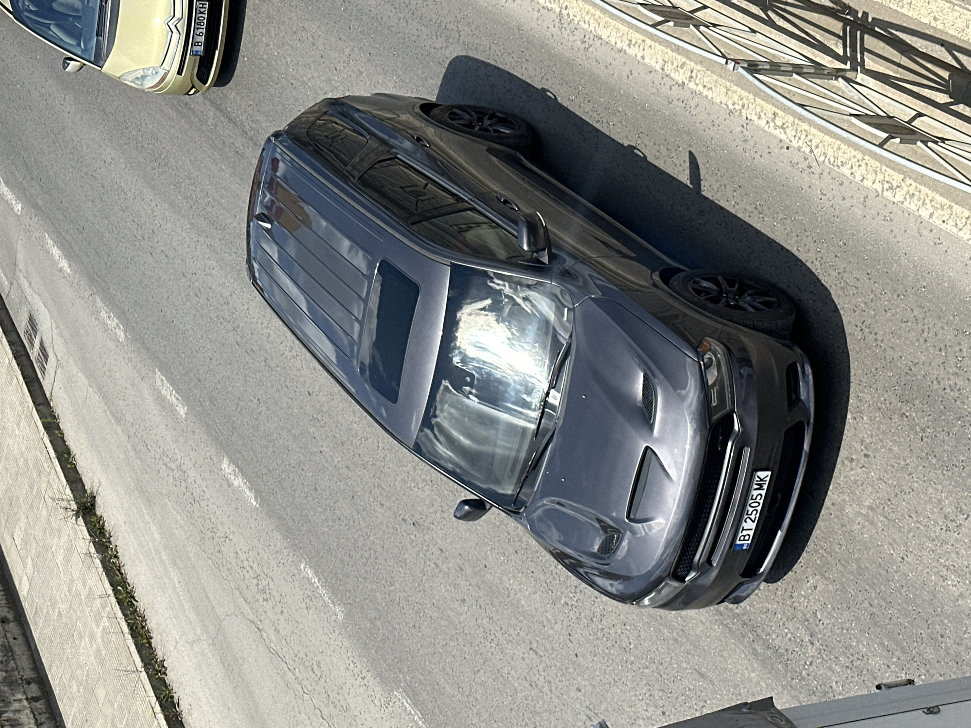 A dark gray Dodge Durango is parked on a paved road in Veliko Tarnovo, Bulgaria. The vehicle has a sunroof and a hood with prominent vents. Its license plate reads "BT 2505 MK". A portion of a yellow Citroen C4 Picasso is visible in the upper right of the frame, with license plate "B 6180 KH". The road has a sidewalk and a metal railing on the right side. The scene appears to be captured from an elevated perspective.
