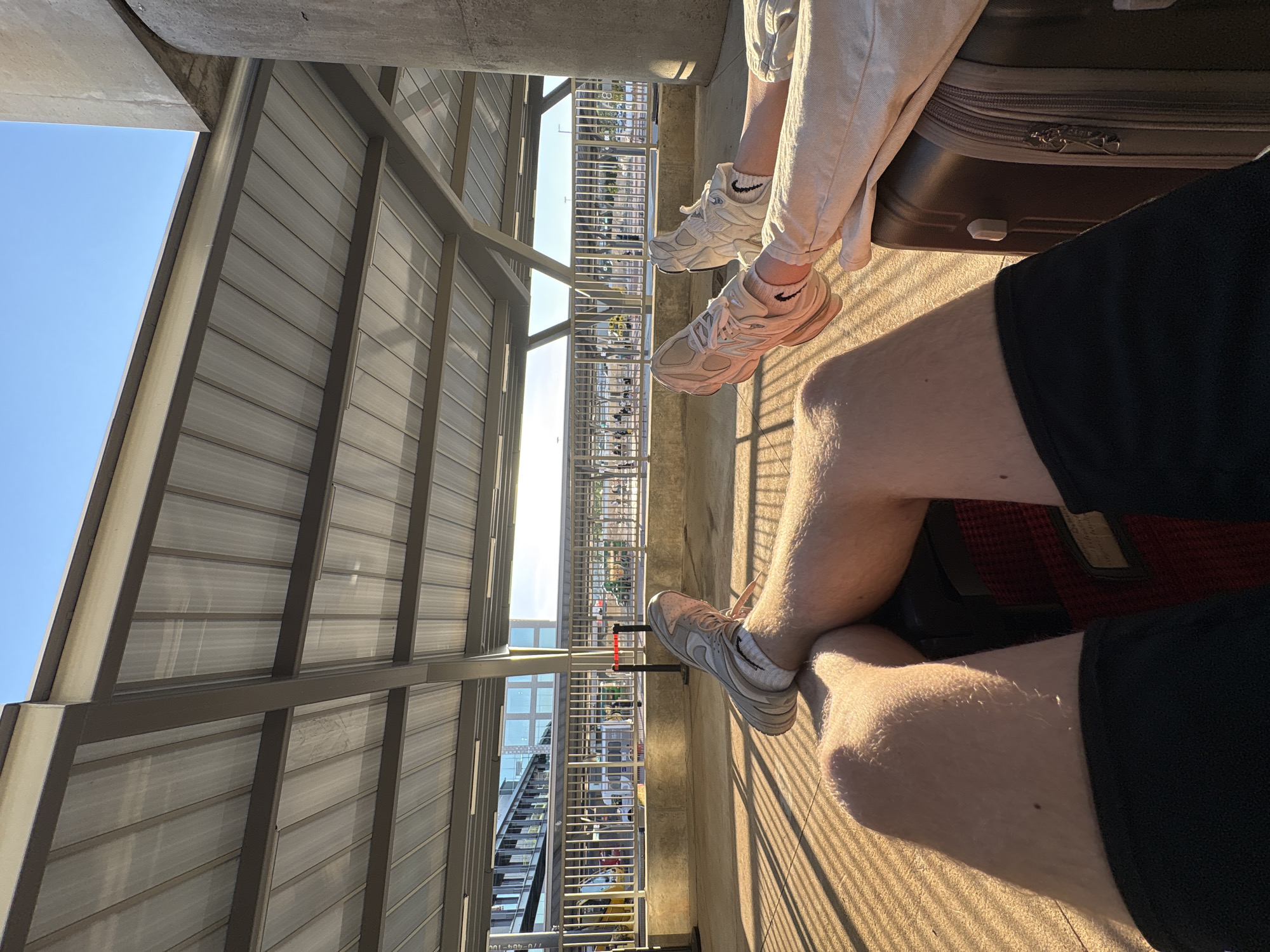 The image presents a first-person perspective of two individuals seated in what appears to be an outdoor area, likely a train station or platform, within College Park, USA. The seating arrangement suggests a journey, with a suitcase partially visible near one of the individuals. The scene is bathed in sunlight, casting shadows across the seating area and legs of the individuals.
One person is wearing light-colored pants and white shoes with a visible black Nike swoosh on their sock. A brown suitcase is adjacent to them. The other person is wearing black shorts and white shoes with a visible black Nike swoosh on their sock.
The backdrop shows a city environment with buildings and infrastructure, viewed through the station's barrier. A roof structure covers the seating area, creating a play of light and shadow.