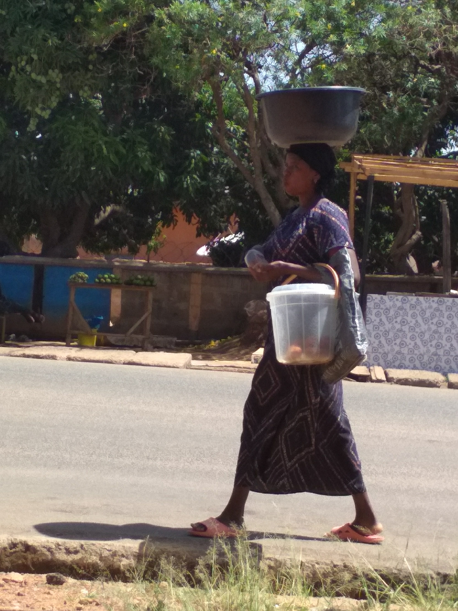 In a street scene in Jos, Nigeria, a woman walks towards the right. She carries a large black basin on her head and a plastic bucket in her left hand. The woman is wearing a long, dark-patterned dress and pink sandals. Behind her is a wooden stand with produce and a tiled wall. Trees line the street, providing shade. The road is paved and stretches out of frame.