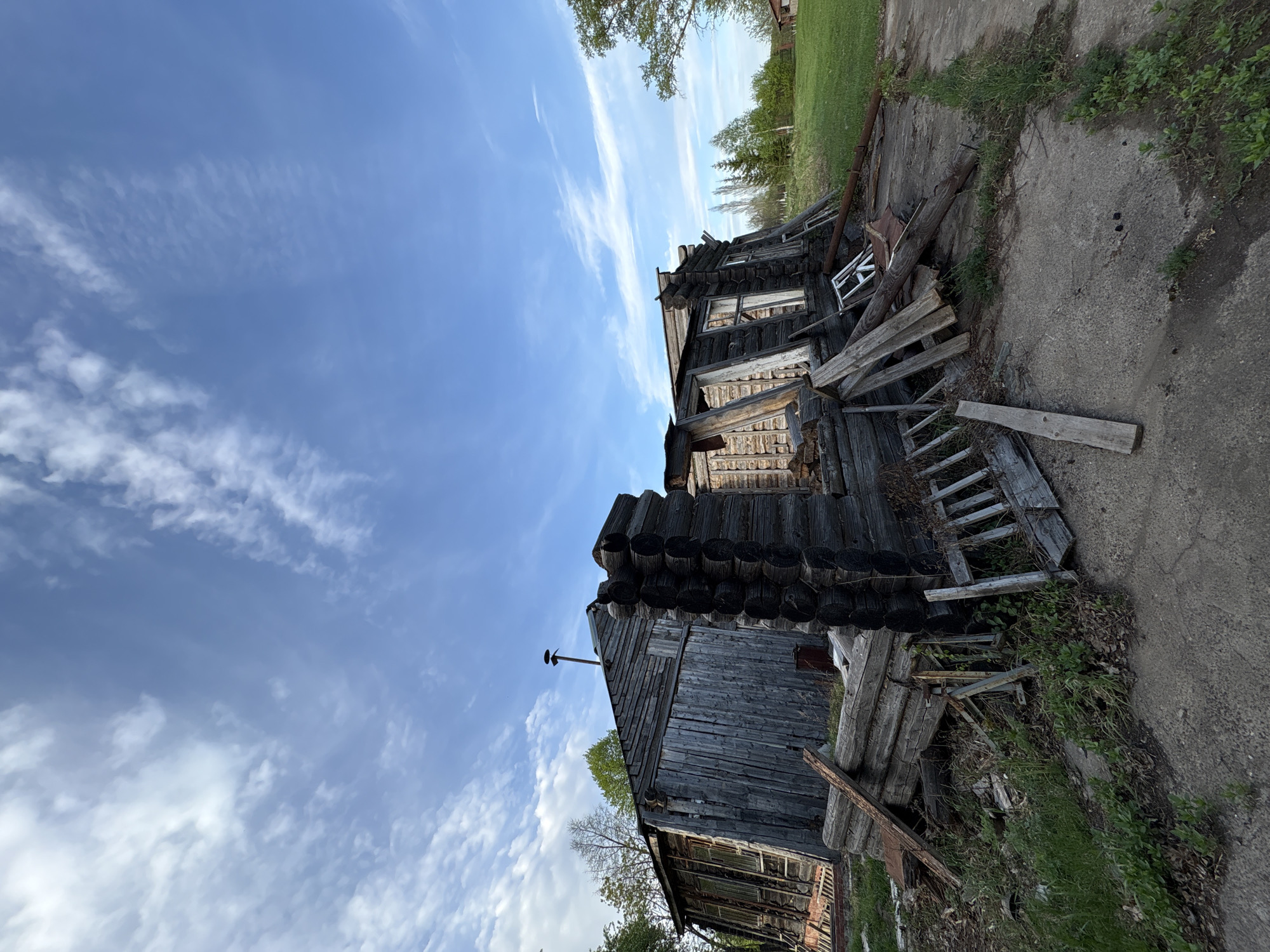 The image depicts a dilapidated wooden building in Zavod Mikhailovskii, Russia. The structure appears abandoned and in disrepair. The building is constructed from logs and weathered wooden planks, with evidence of decay and structural damage. Debris, including wooden boards and other materials, are scattered around the base of the building. A concrete or paved area surrounds the building, with vegetation growing through cracks and along the edges. The sky above is blue with wispy clouds.