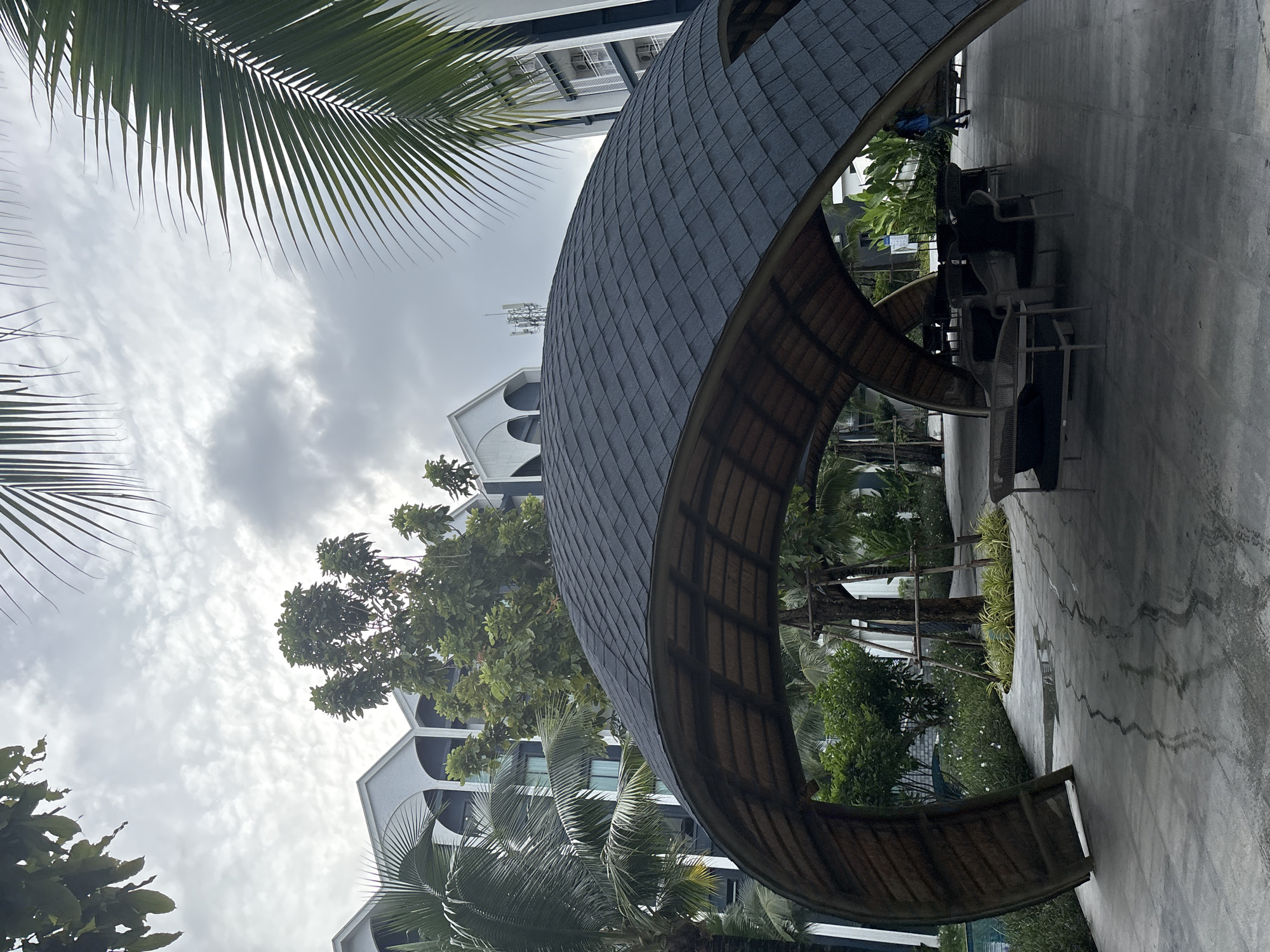 A person walks past a covered outdoor seating area with curved, dark gray shingled roofs supported by bamboo-like structures, near a resort in Sakhu, Thailand. The scene depicts an overcast day, with palm fronds framing the top of the image. In the background, multi-story resort buildings with arched windows are visible, surrounded by lush green trees and plants. The ground is paved with concrete, showing water stains and patterns. The seating area includes dark upholstered lounge furniture.