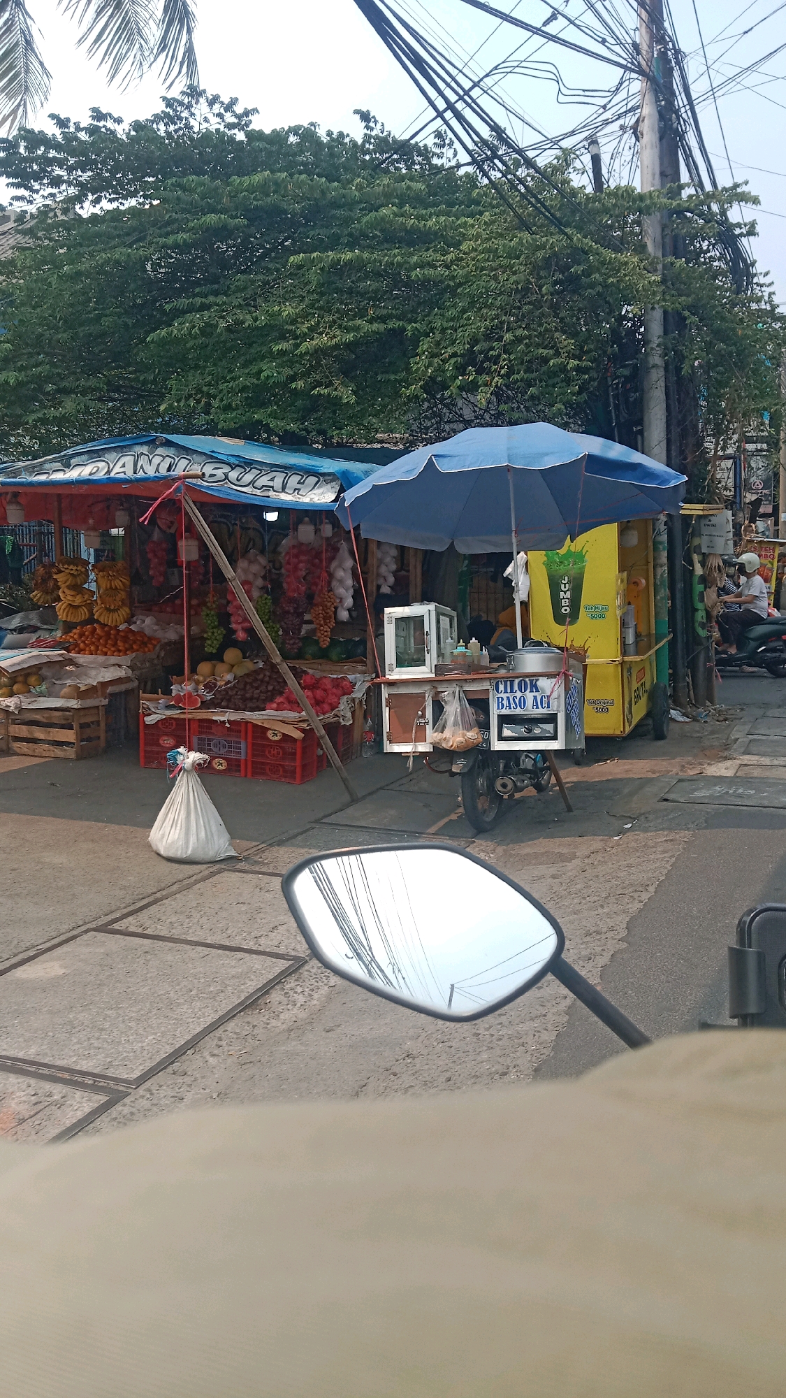 The image captures a street scene in Bekasi, Indonesia, where a fruit stall and a food cart are set up along the roadside. The fruit stall displays a variety of fruits, including bananas, oranges, grapes, and what appear to be rambutan or lychee. Several crates and wooden boxes are used to present the produce. Behind the fruit stall, lush green trees provide a natural backdrop.
Adjacent to the fruit stall is a yellow food cart, distinguished by a blue umbrella overhead. The cart is equipped with a display case and signage that reads "CILOK BASO ACI." A plastic bag filled with what looks like bread or snacks hangs from the cart. A motorcycle is parked at the cart, suggesting it's part of the mobile food operation. Further in the background, utility poles with a dense network of wires are visible, a common sight in urban Indonesian environments. A person is seen sitting on a motorcycle in the far right of the image, with another individual standing nearby. The ground is paved, with some square concrete slabs and asphalt. A white sack is placed on the ground in front of the fruit stall. The reflection in a motorcycle's side mirror in the foreground shows a portion of the scene and overhead wires.